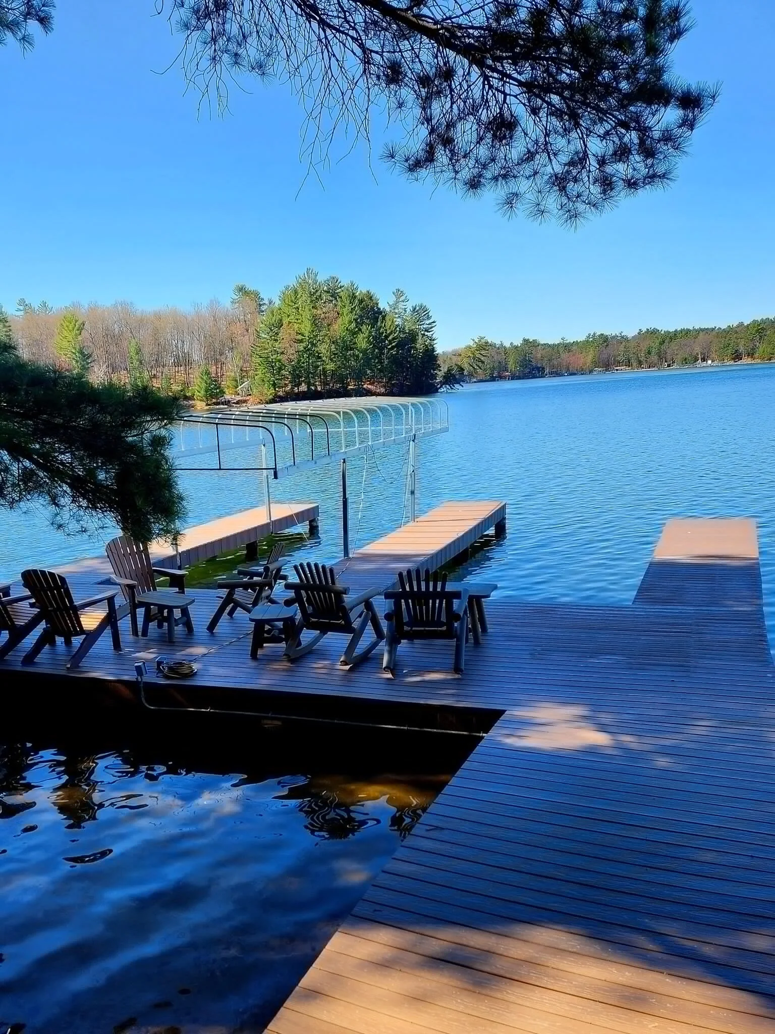 A lakeside dock with Adirondack chairs, wooden planks, and a boat lift on a calm lake with a tree-lined shore and blue sky.