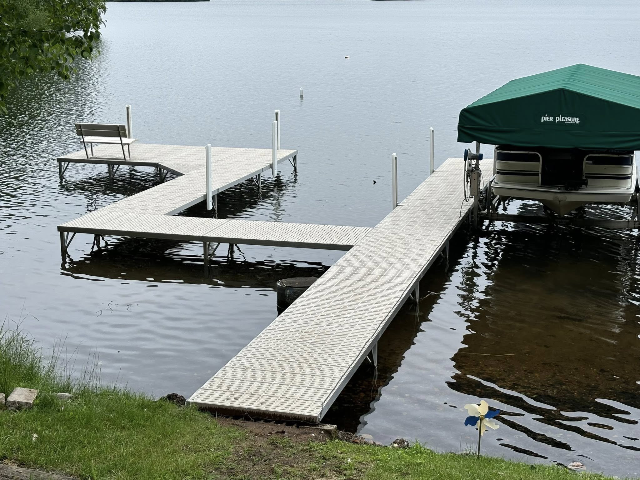 A metal boat dock extending into a calm body of water, with a small seating area at the end, and a covered boat lift on the right side of the dock, surrounded by green grass and a single white flower in the foreground.