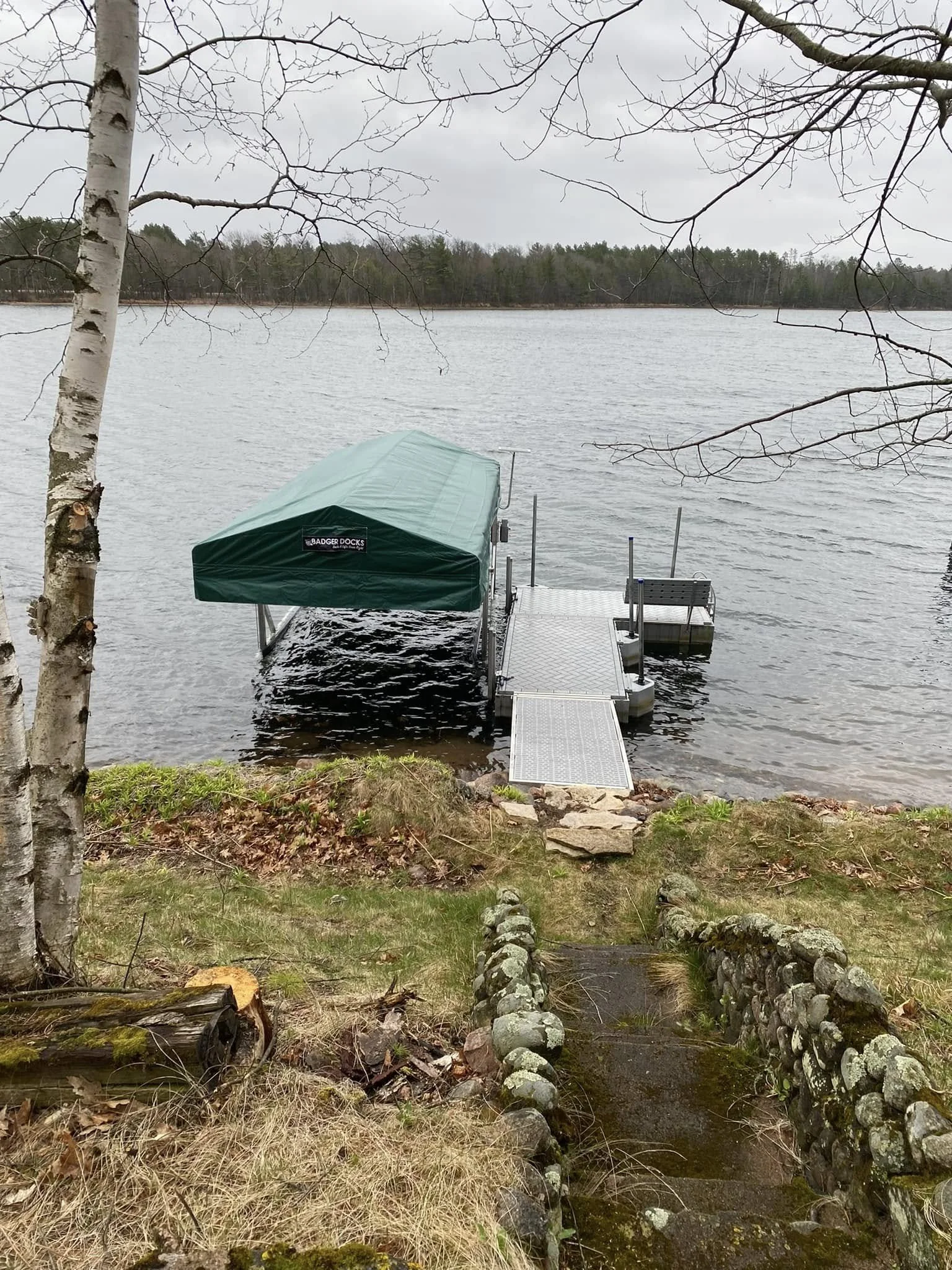 A lakeside scene with a small dock extending into water, featuring a covered boat slip and floating platform. The foreground shows stone steps and a grassy area with a leafless tree on the left.