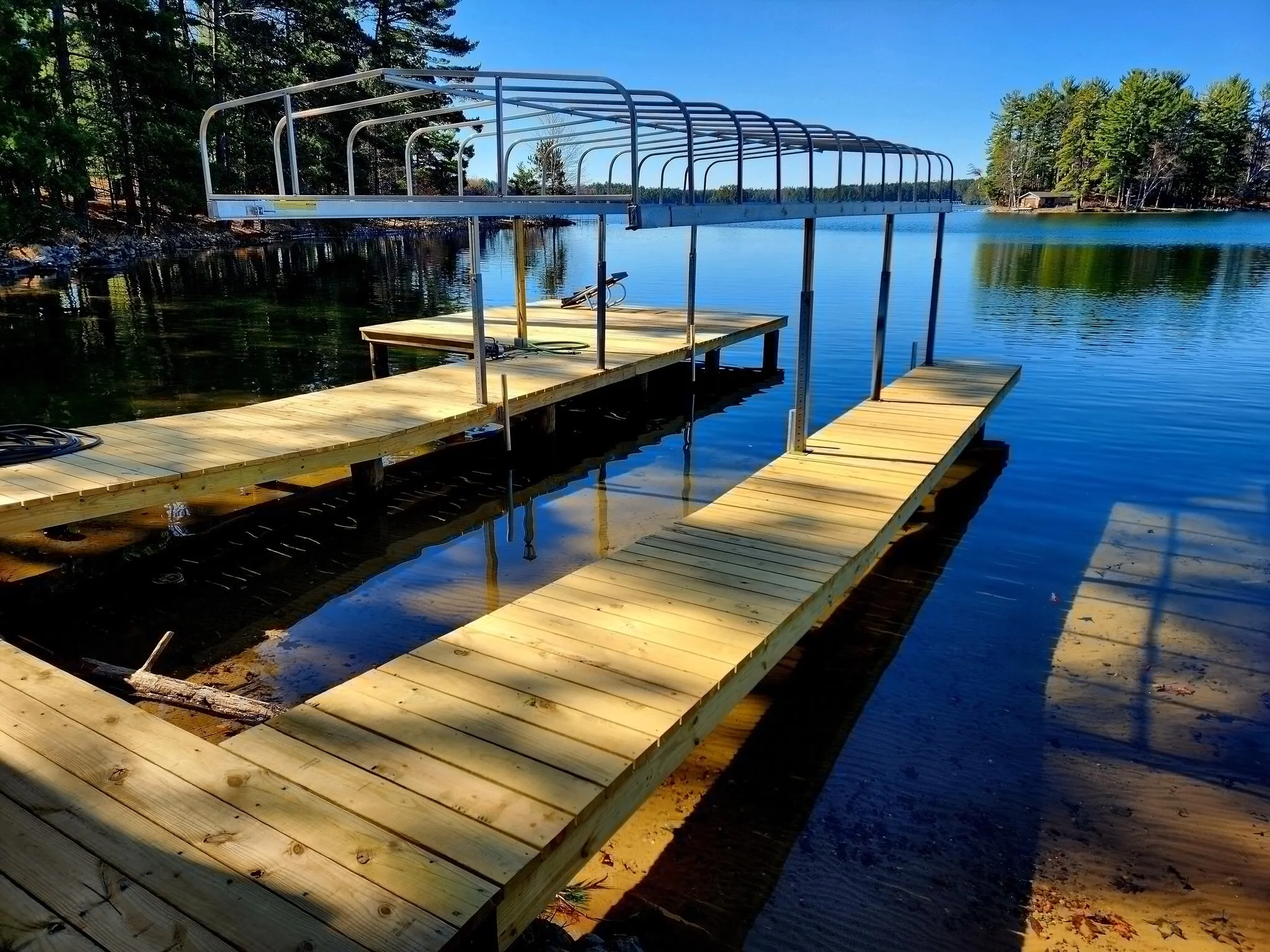 Wooden dock with a metal boat lift on a calm lake, surrounded by trees and a clear blue sky.