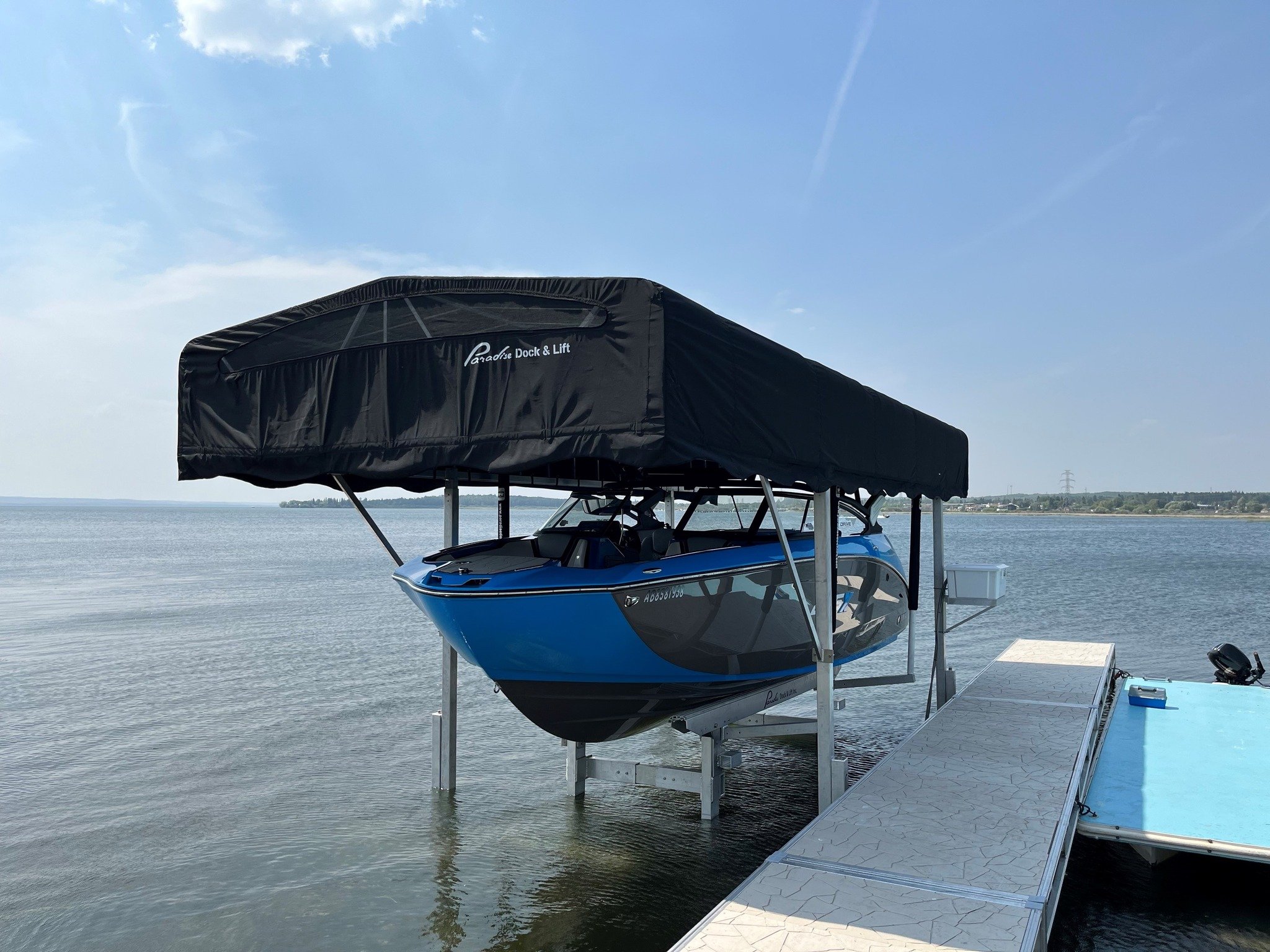 A boat lift holding a blue and black boat with a black canopy on a calm body of water, with a distant shoreline and clear sky.