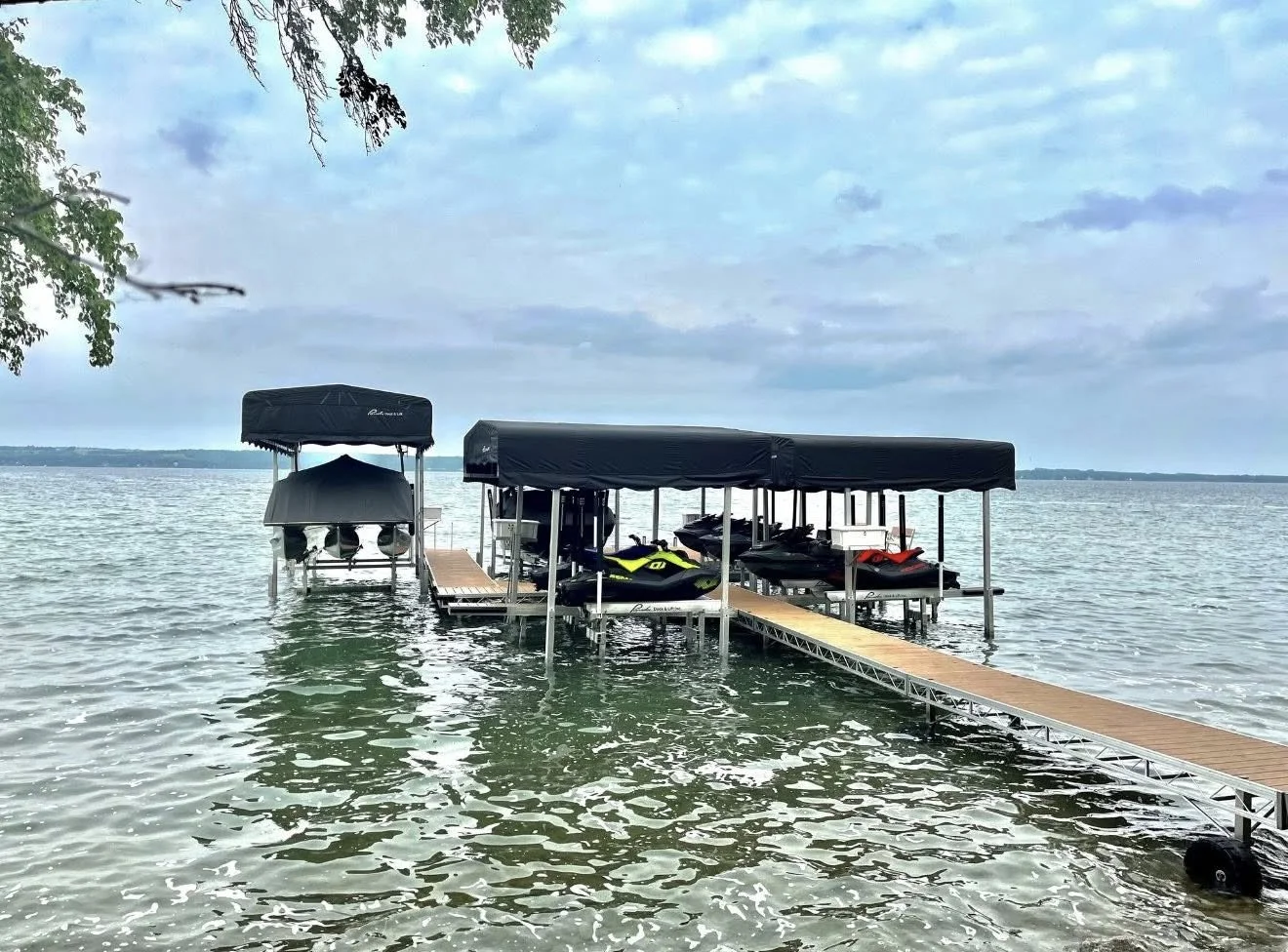 A boat dock with canopies, holding two jet skis and other watercraft, extends into a large lake under a cloudy sky.