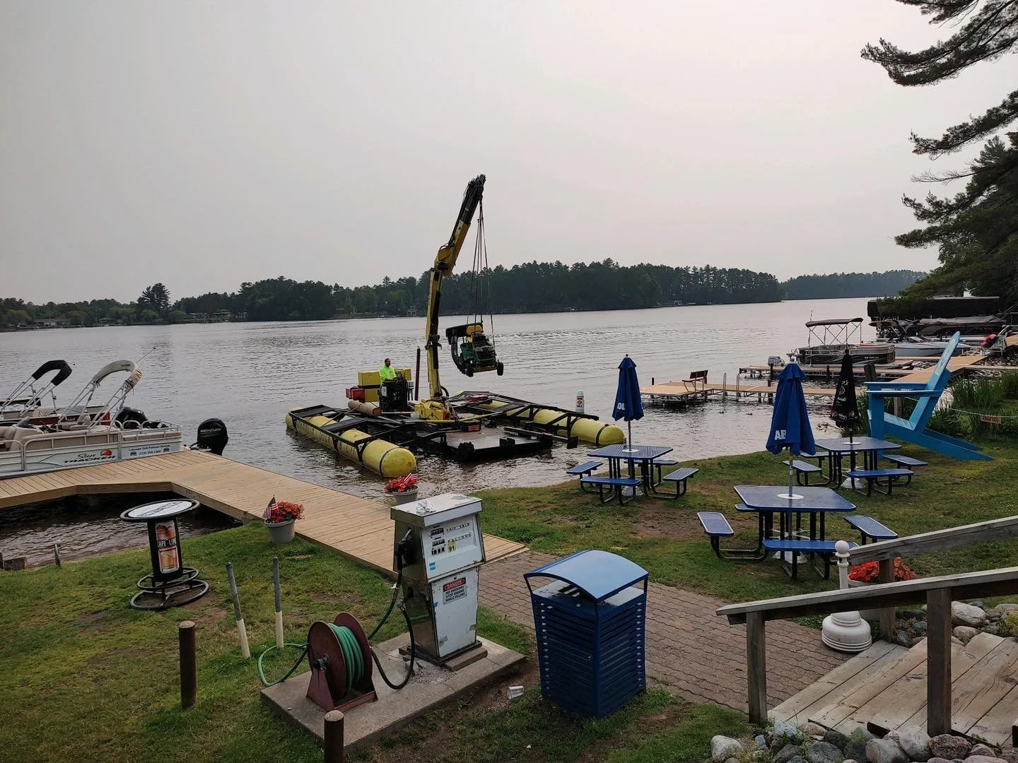 A lakeside scene with a dock, boats, and outdoor seating areas. A boat lift with a crane is positioned in the water. Several boats are docked along the shore. There are tables with blue umbrellas on a grassy area, and a trash can, a hose reel, and re