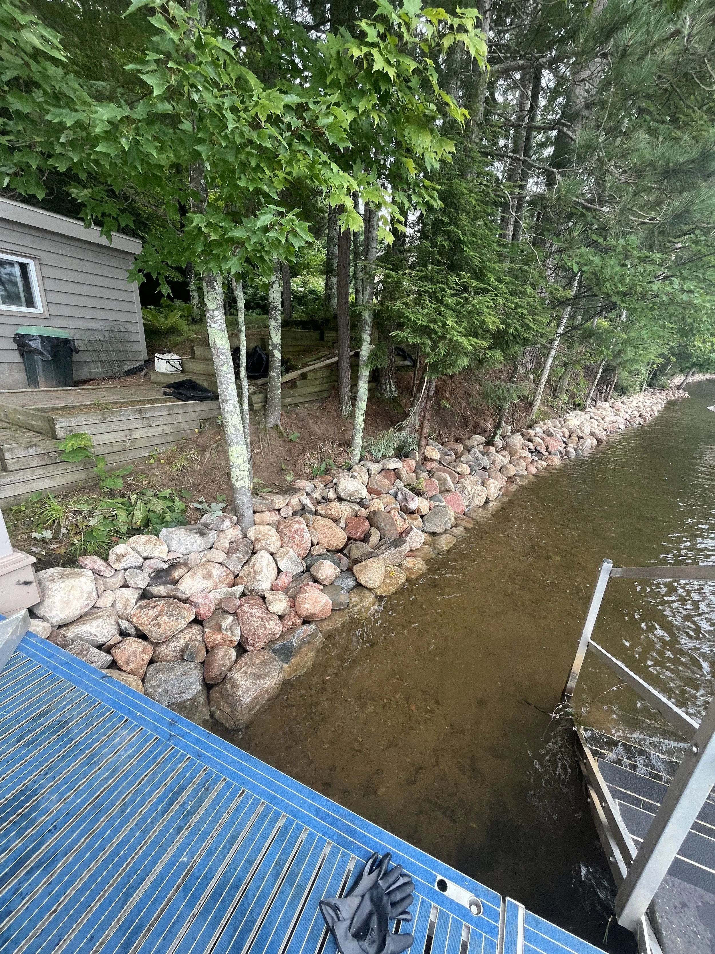 View of a lakeside shoreline with a wooden dock, a set of metal stairs leading into the water, rocks lining the shoreline, and a house partially visible among trees.