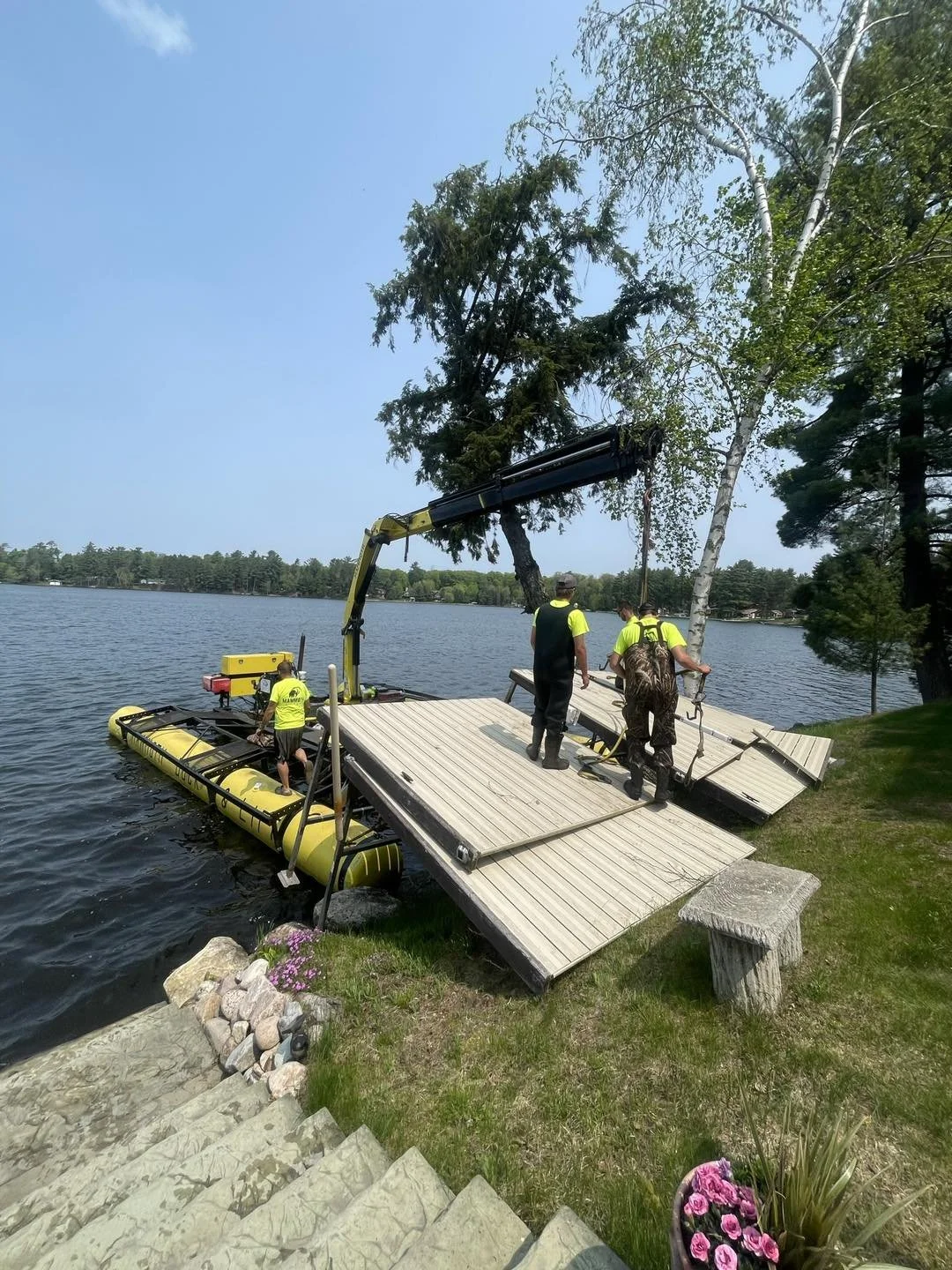People launching a yellow pontoon boat from a lakeside dock on a sunny day, with trees in the background.