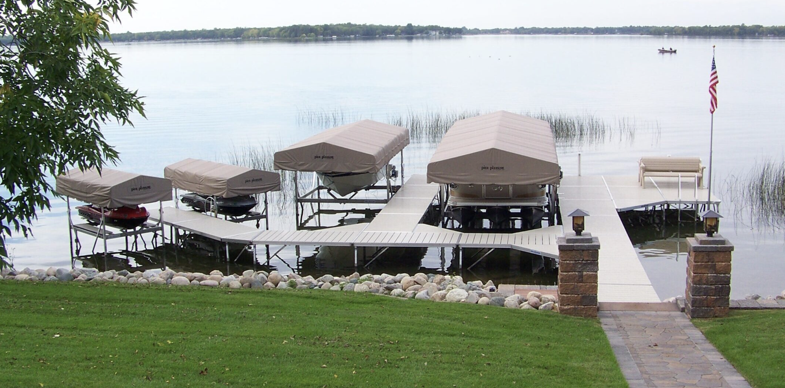 A dock with boats and boat covers on a lake, with a grassy area and stone pathway in the foreground, and an American flag on the right.