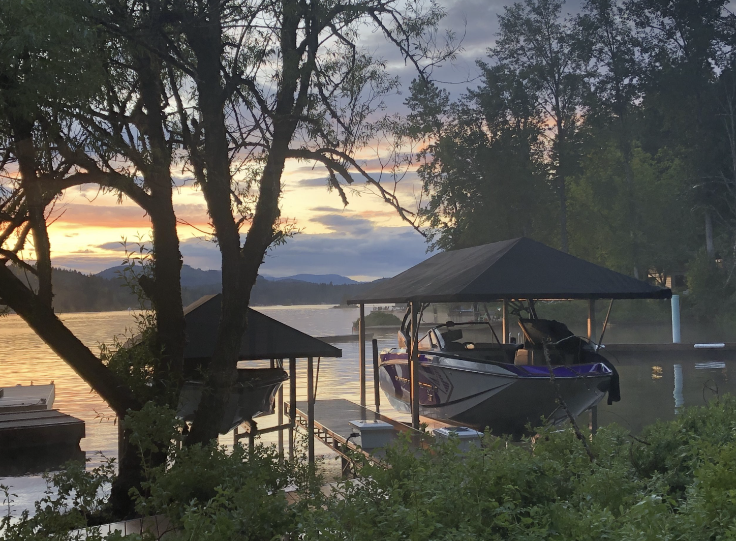 A boat docked at a lake during sunset, surrounded by trees and mountains in the background, with calm water and a partly cloudy sky.