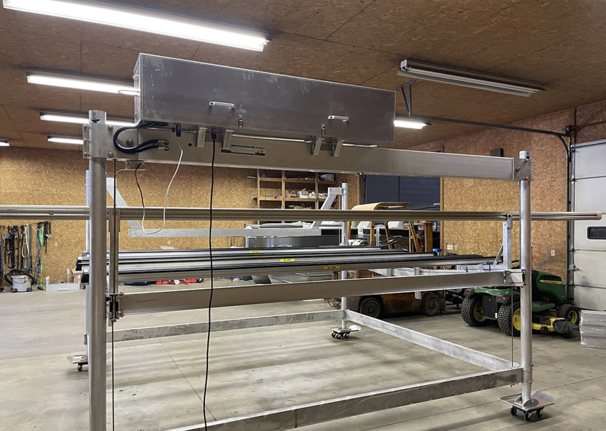 A large empty metal frame structure on wheels in a workshop or garage, with a metallic box attached at the top and some wires hanging from it, surrounded by various tools and equipment.