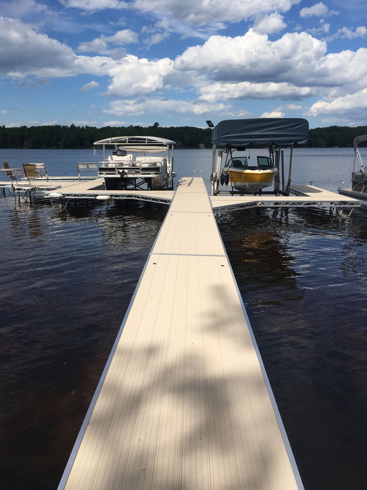 A dock extending into a lake with three boats tied to it, under a partly cloudy sky, with trees in the background.