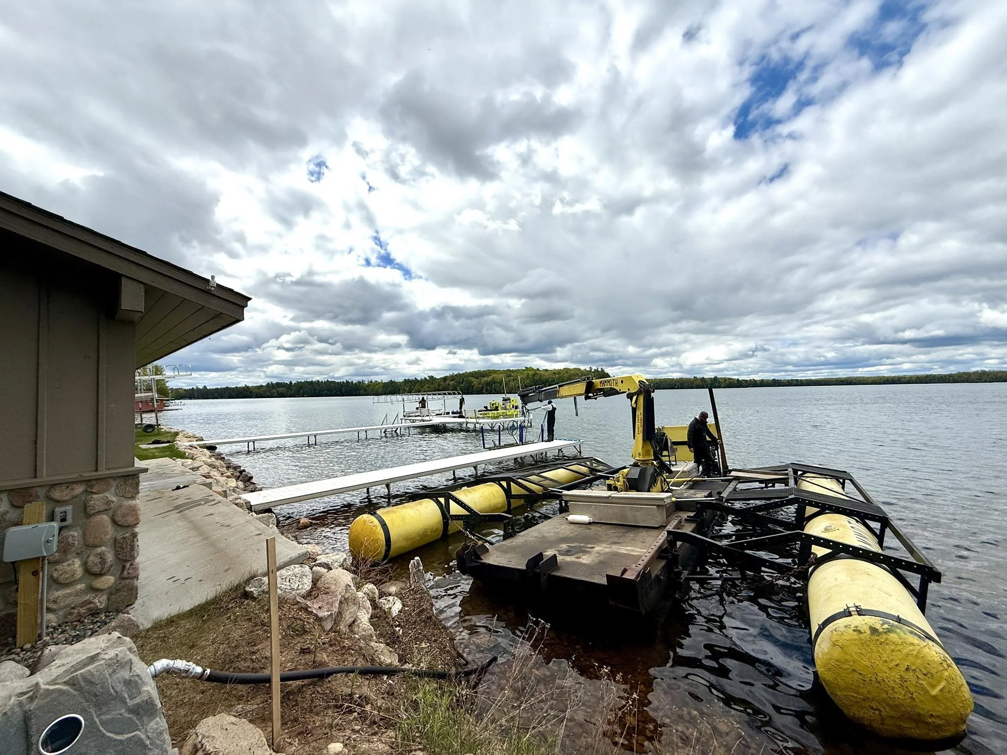 Industrial watercraft being assembled or repaired on a lakeshore, with cloudy sky and other boats in the background.
