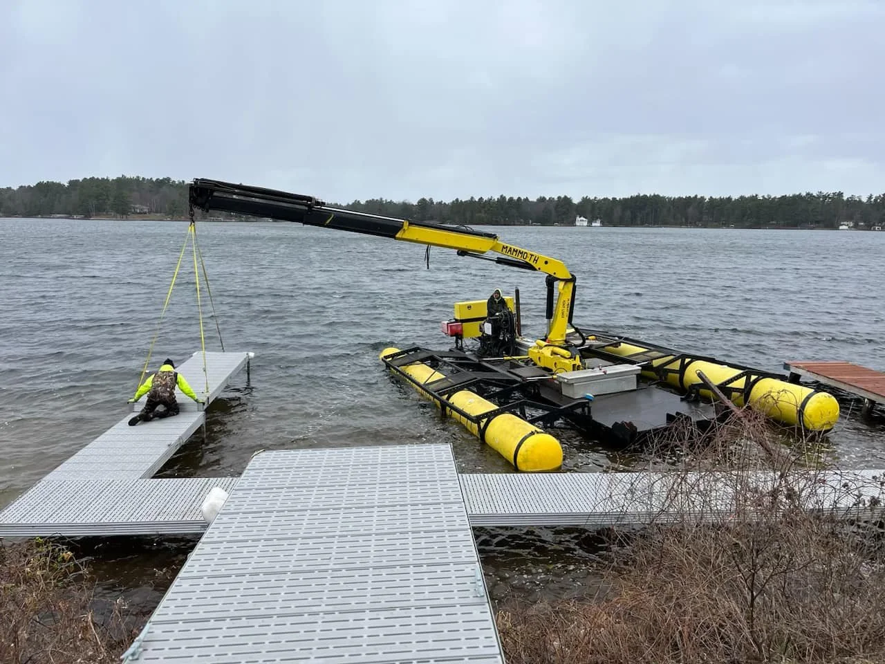 A drone boat with yellow pontoons and a crane is lifting a metal dock panel into a body of water on a cloudy day.