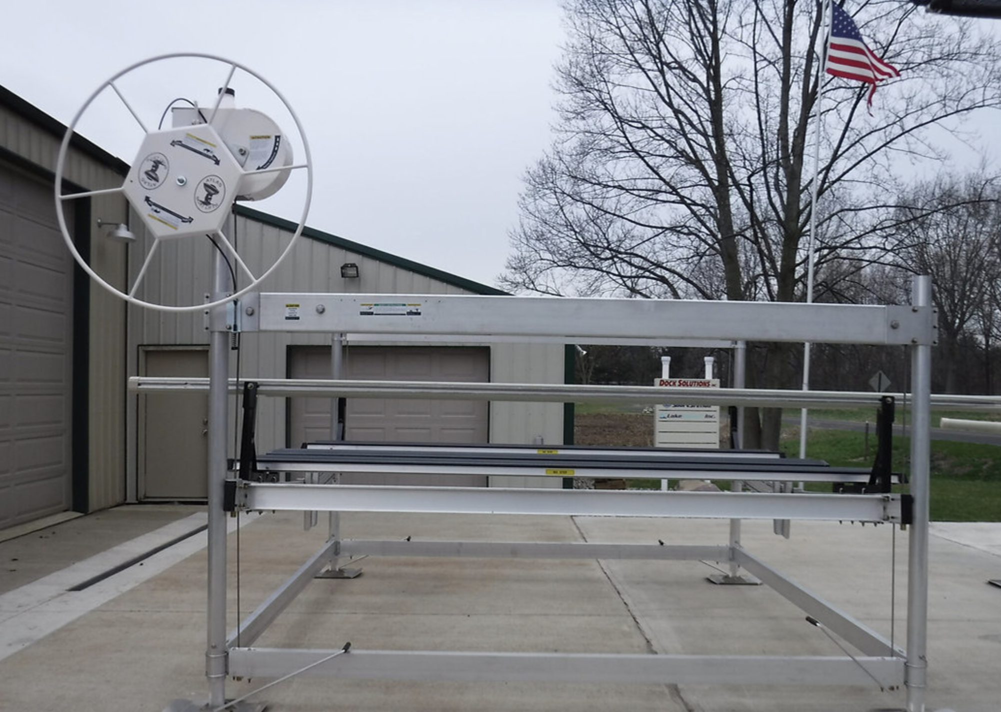 A metal dock lift platform with a rotating wheel and an American flag in the background, set outdoors on a concrete surface near a storage building, with trees and a cloudy sky.