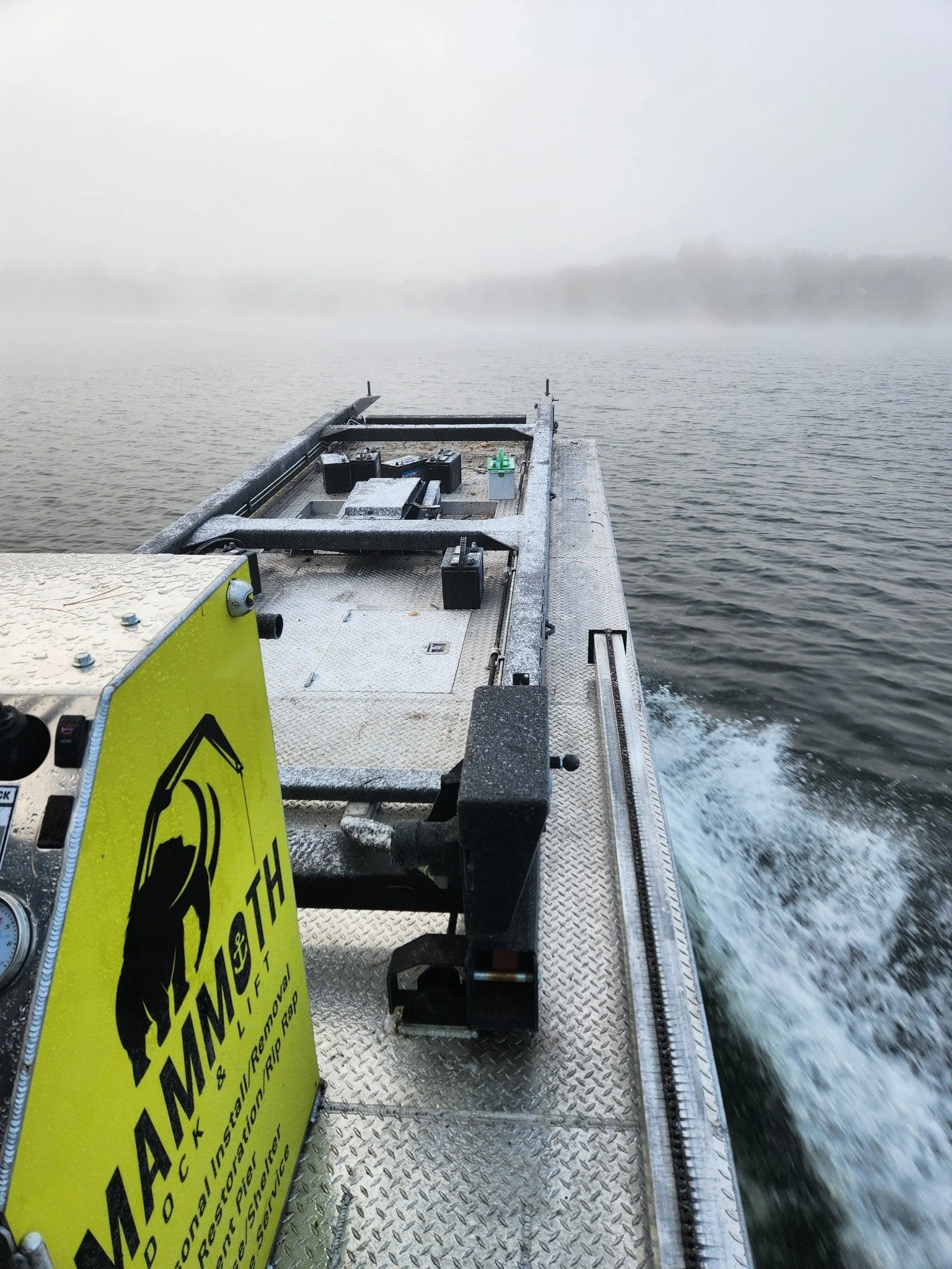 View from the front of a boat moving on a body of water with foggy weather and trees in the distance; the boat's deck has equipment and a bright yellow warning sign that reads 'WARNING' with symbols of a person falling and a message about slip and trip hazard.