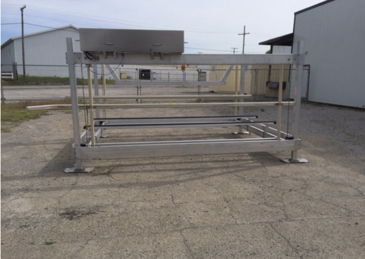 An empty, outdoor, industrial metal platform with rails and a control box, situated on a cracked asphalt surface, with industrial buildings and utility poles in the background.