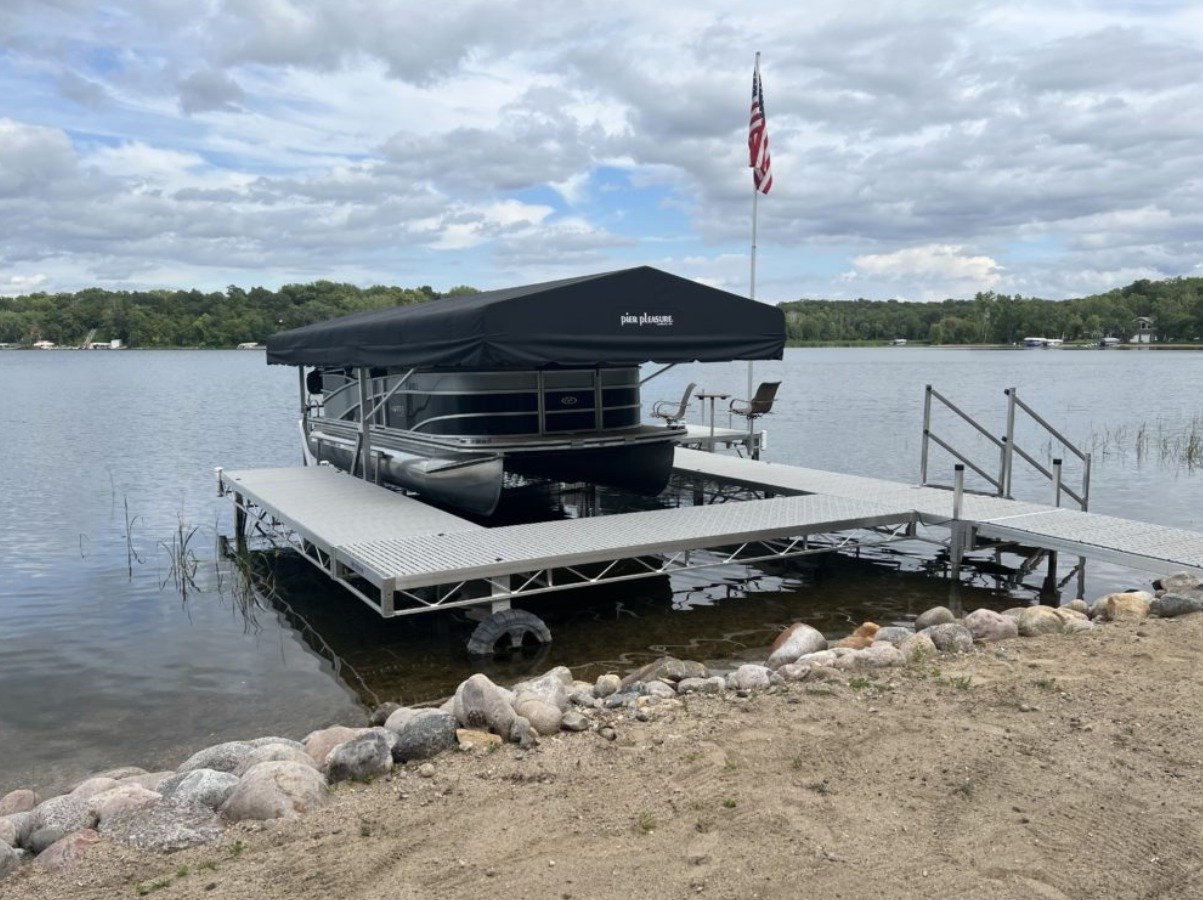 A pontoon boat docked at a lakeside with a covered canopy and two chairs, surrounded by water, rocks, and a cloudy sky.