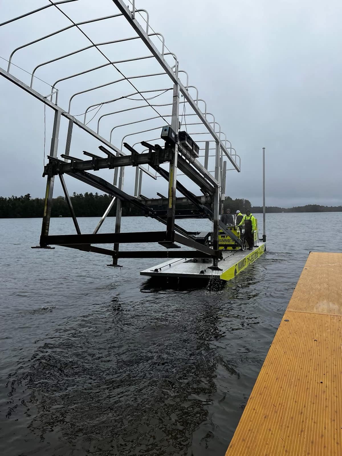 A boat docked at a pier with a metal structure and safety railing, two workers in yellow safety vests on board, over a body of water on a cloudy day.