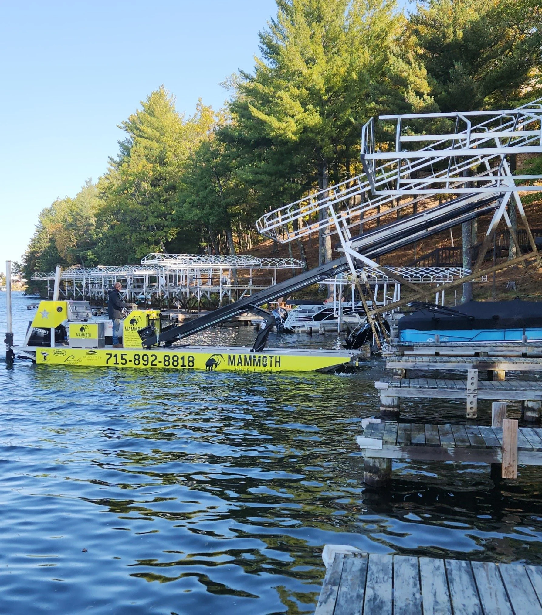 A yellow boat with the words 'Mammoth' and a phone number is floating near a dock. A large, dismantled amusement park ride is on the dock and extending into the water. Green trees and a clear blue sky are in the background.