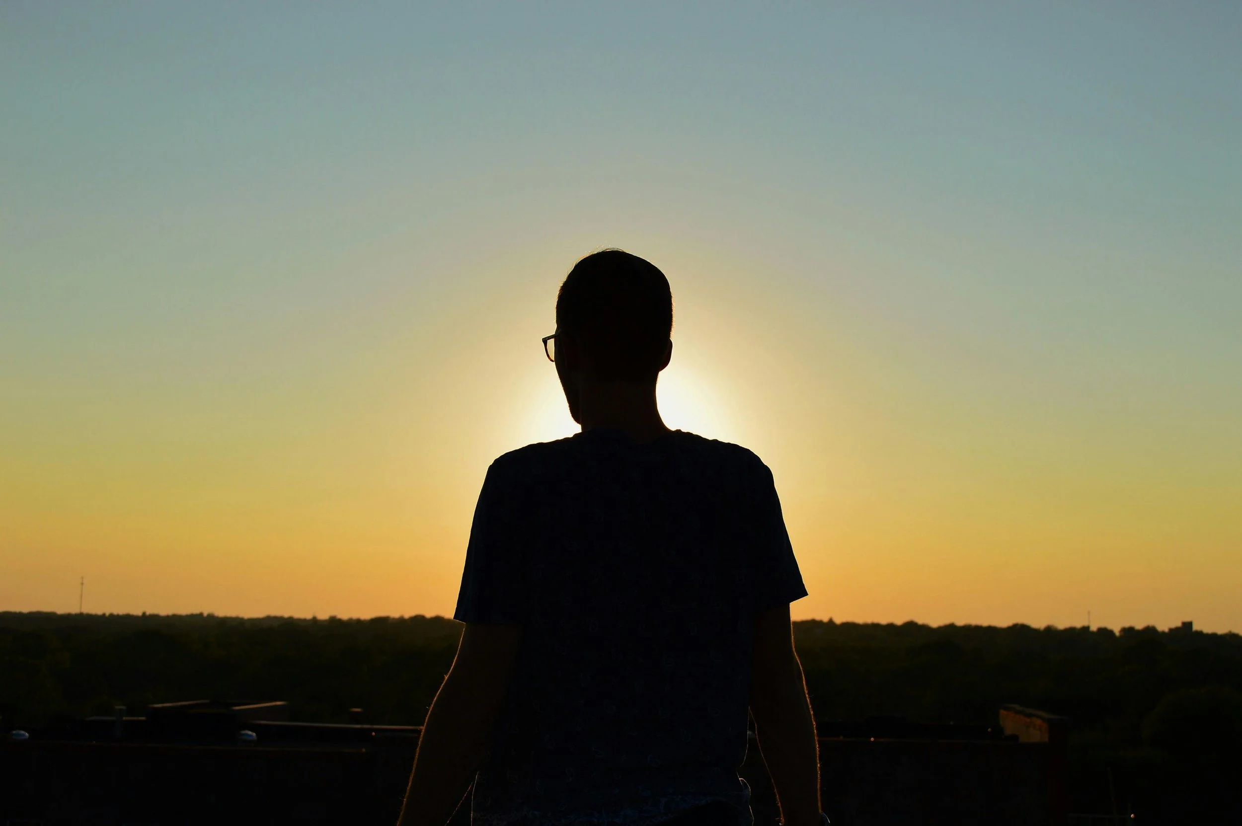 Silhouette of a person wearing glasses standing outdoors during sunset or sunrise.