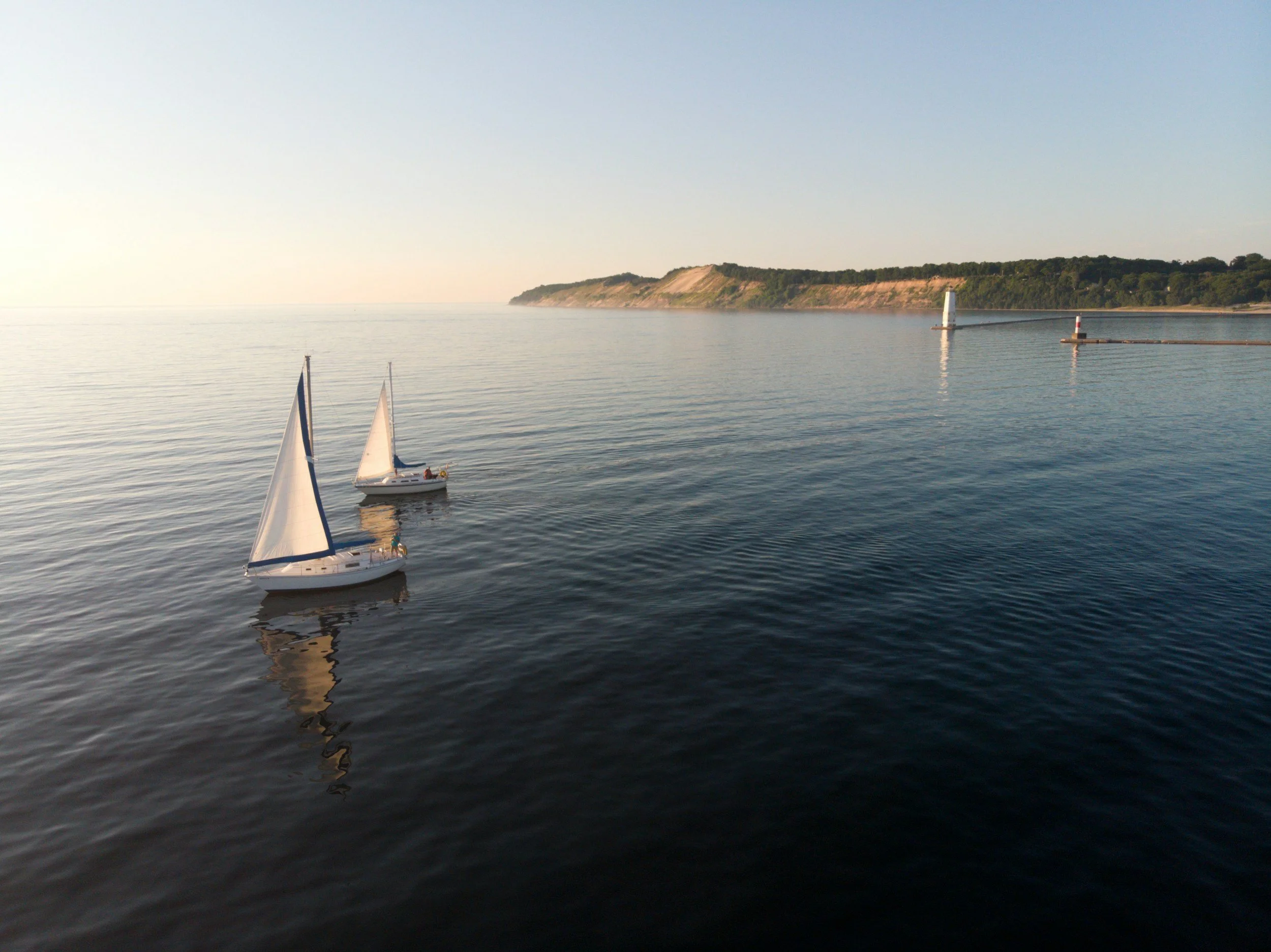 Two small sailboats floating on calm water near a shoreline with green cliffs and two lighthouses in the distance during sunset or sunrise