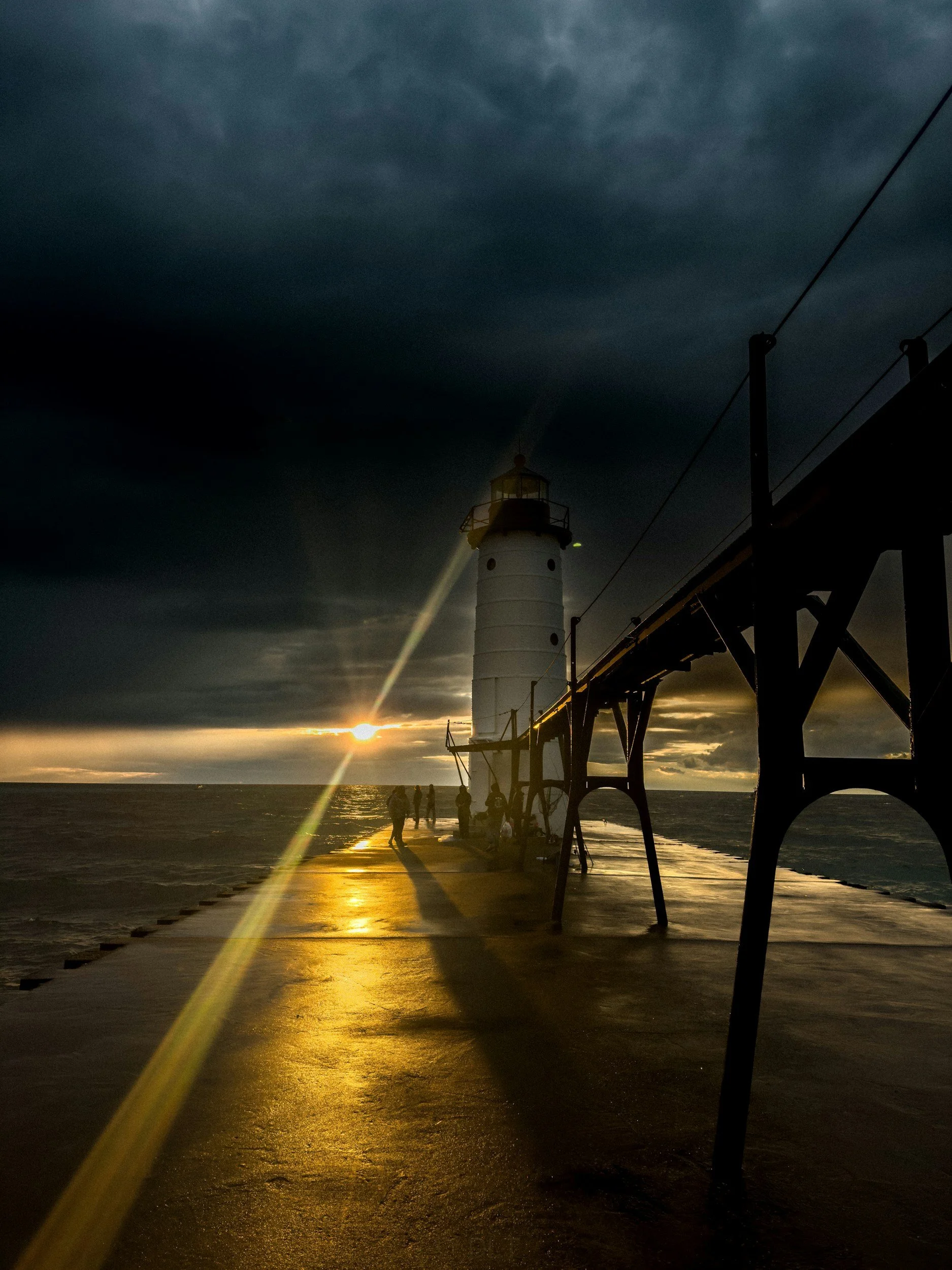 A lighthouse on a pier at sunset, with clouds overhead and a few people walking along the pier.