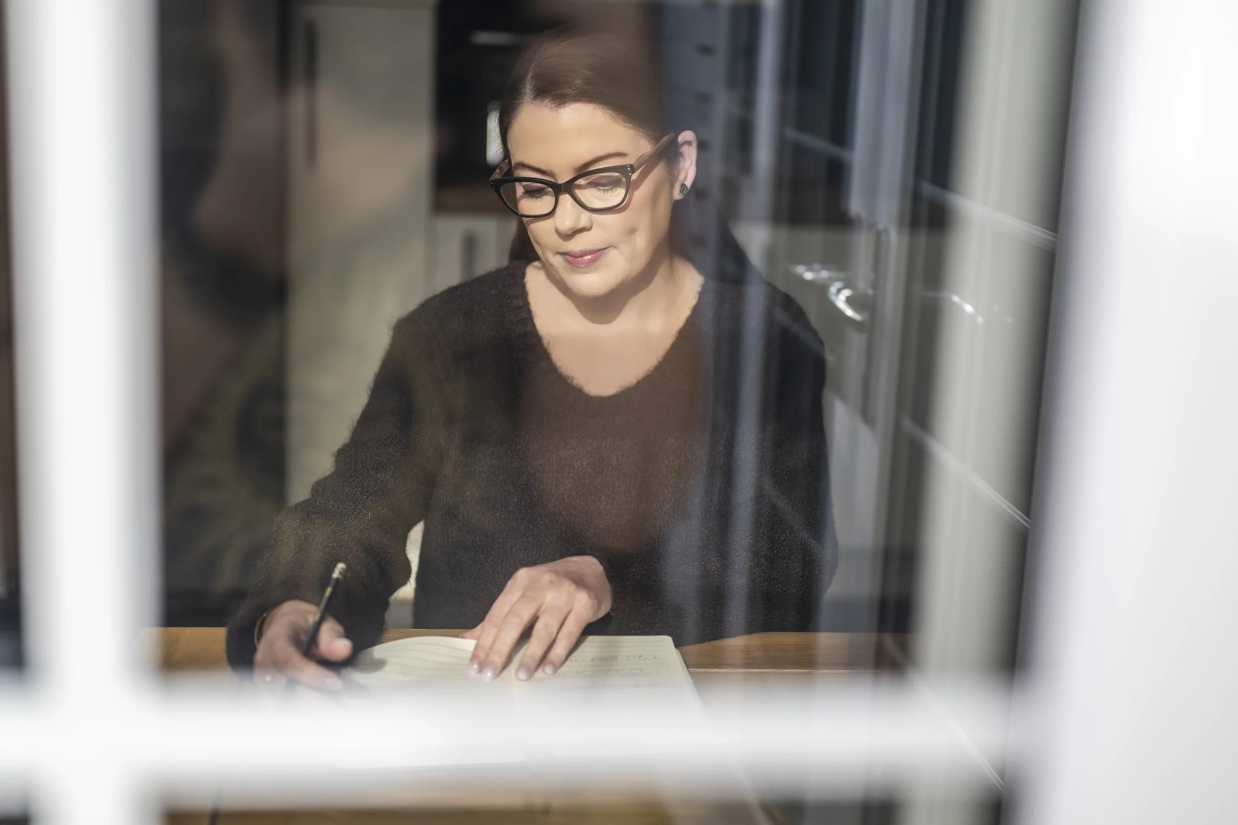 A woman with glasses and dark hair writing in a notebook at a wooden table, seen through a glass window.