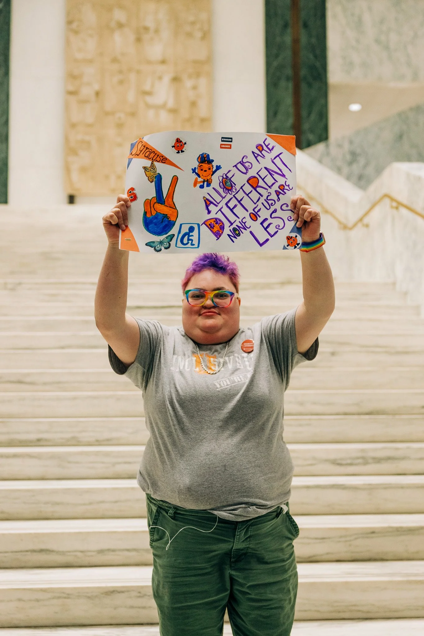 Self Advocate holding up a sign on the steps of the Capital that says "All of us are different, none of us are less."