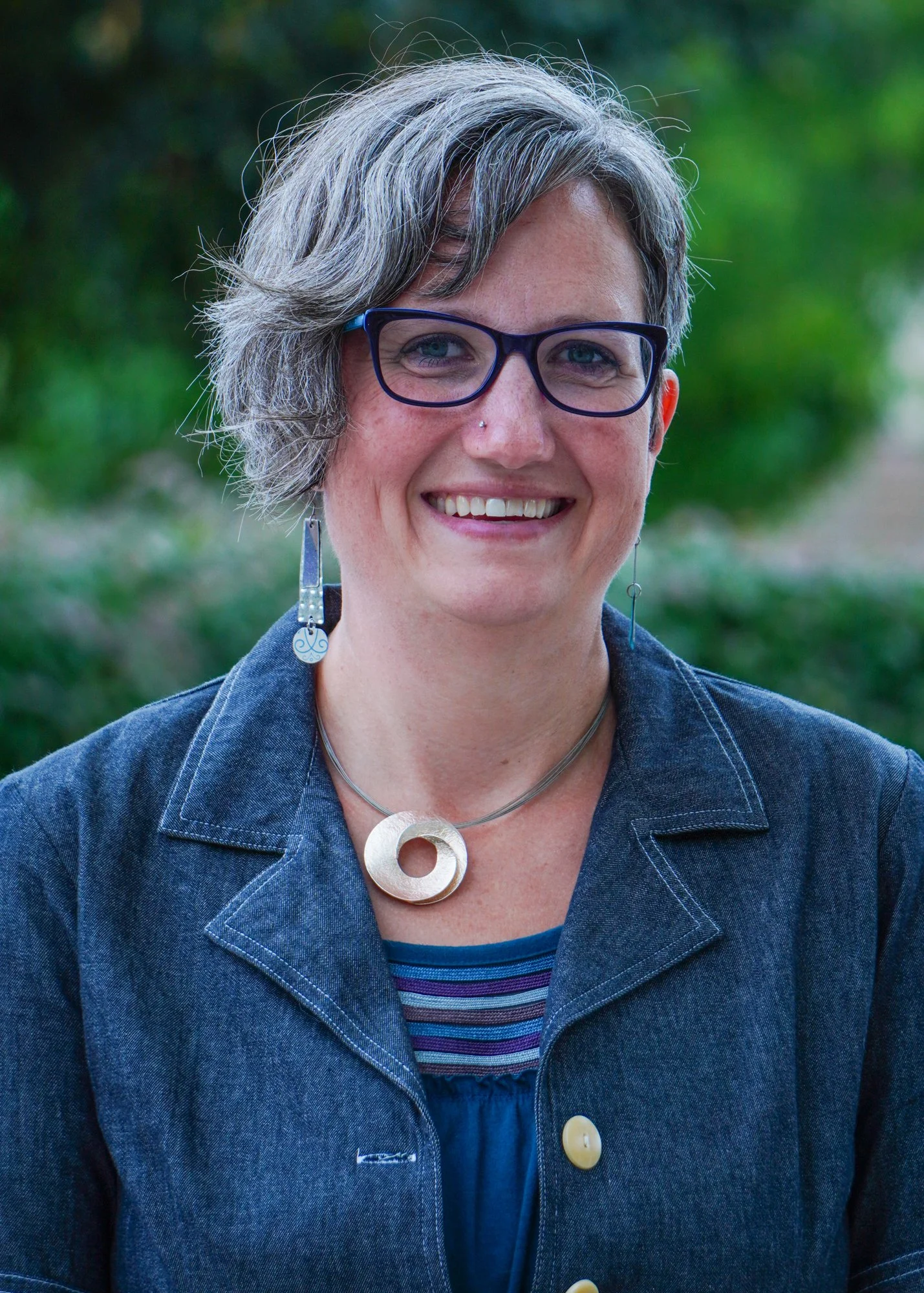 Photo of Sara Petit-McClure, Caucasian woman with short gray hair and glasses with blue framed, chunky jewelry and a jean jacket