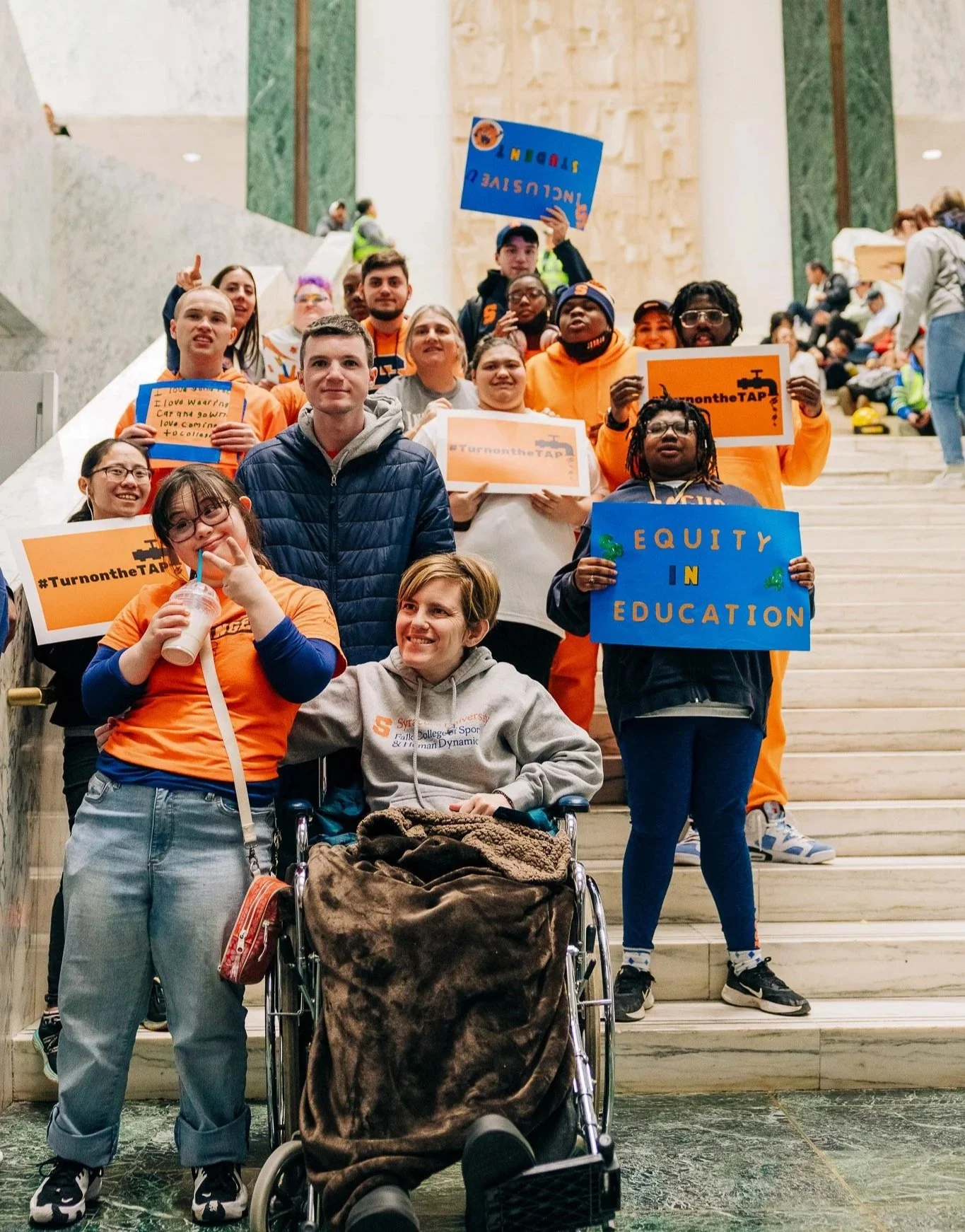 Group of diverse young adults gathering on the steps of a government building, holding signs that promote equity in education and support for the community, with some wearing orange shirts and others in casual attire.