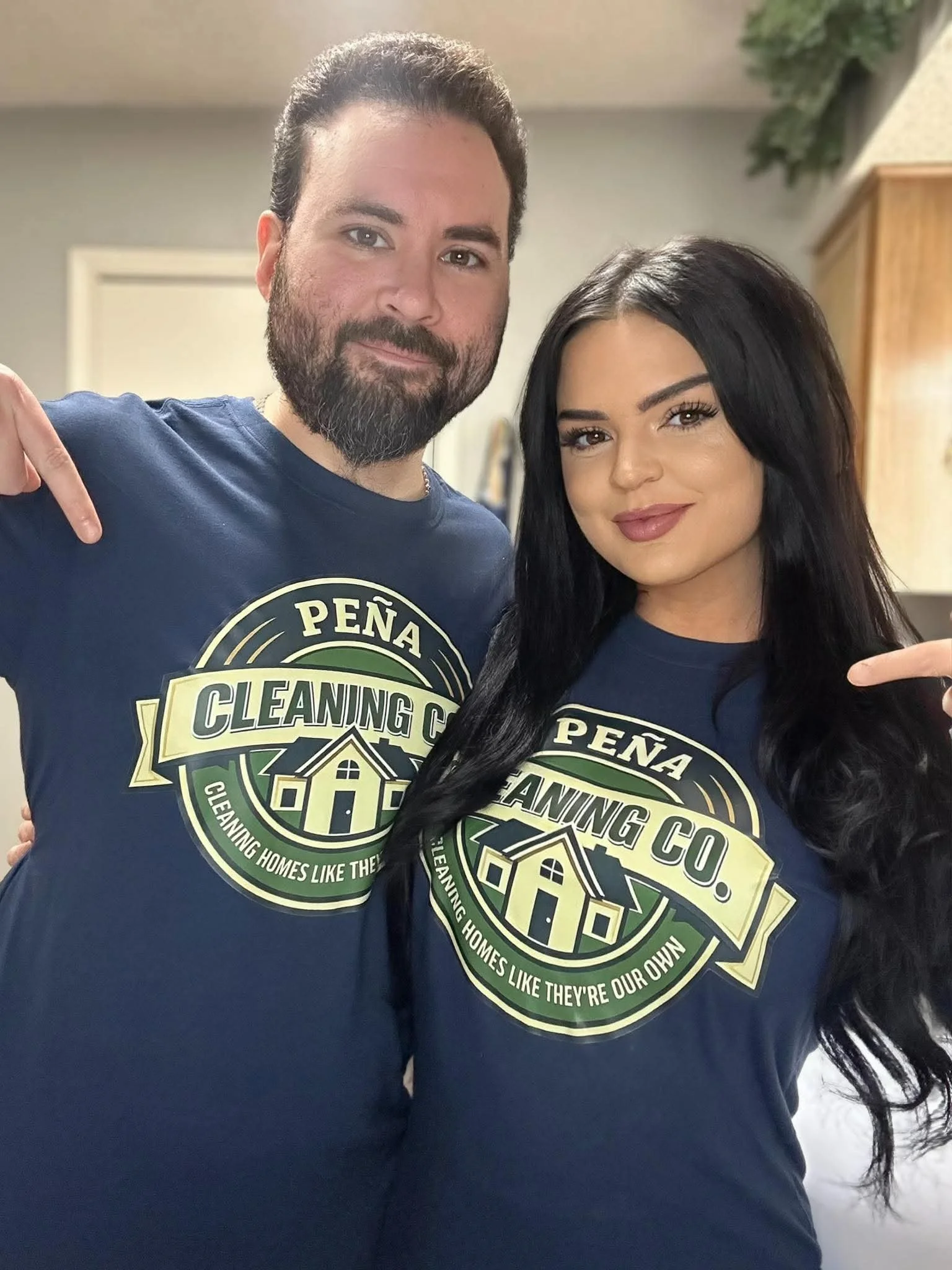 Two individuals wearing matching dark blue T-shirts with the logo of Pena Cleaning Co., pointing at their shirts, inside a room with neutral walls and wood cabinets in the background.