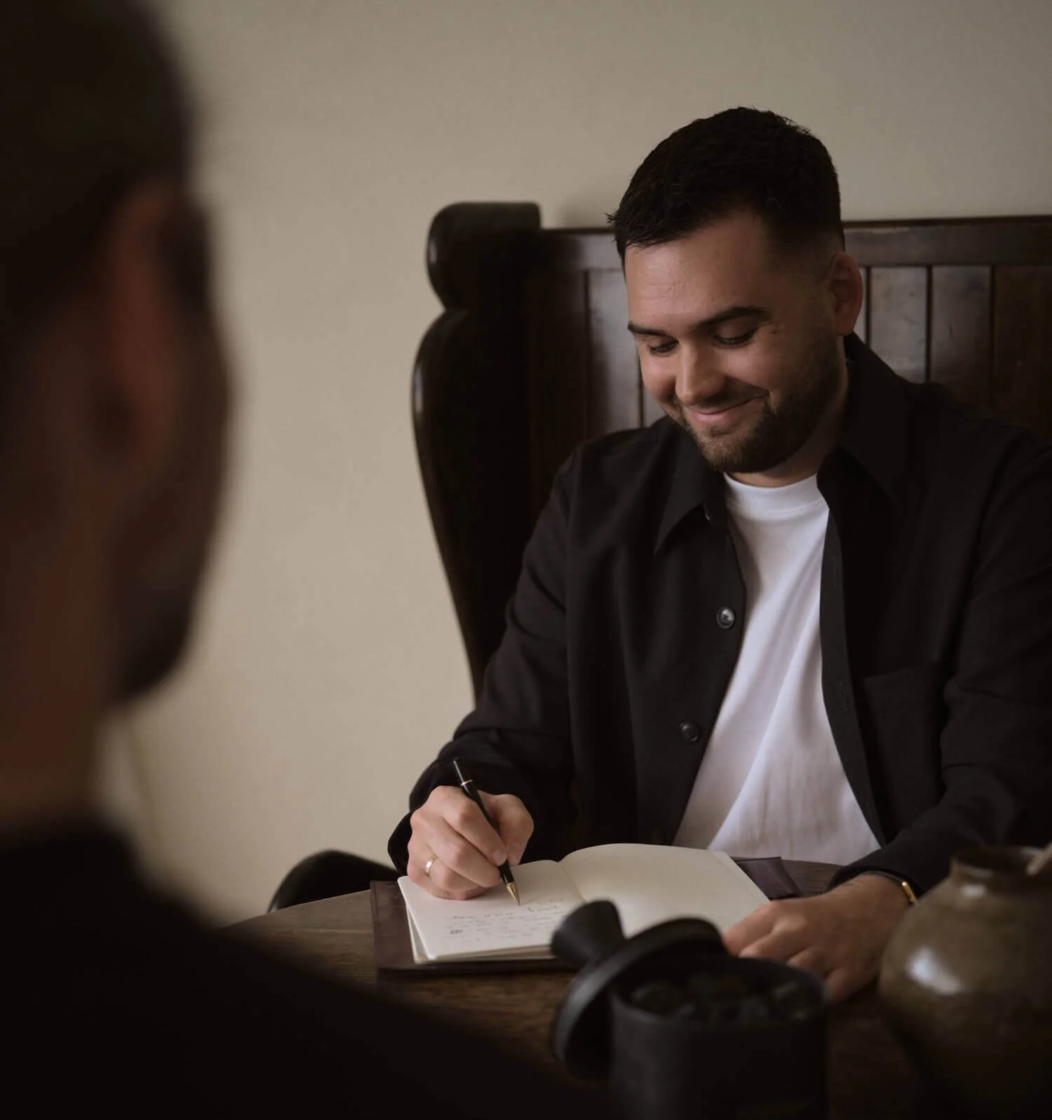 Un homme assis à une table, écrivant dans un carnet, souriant, avec une autre personne légèrement floue au premier plan.