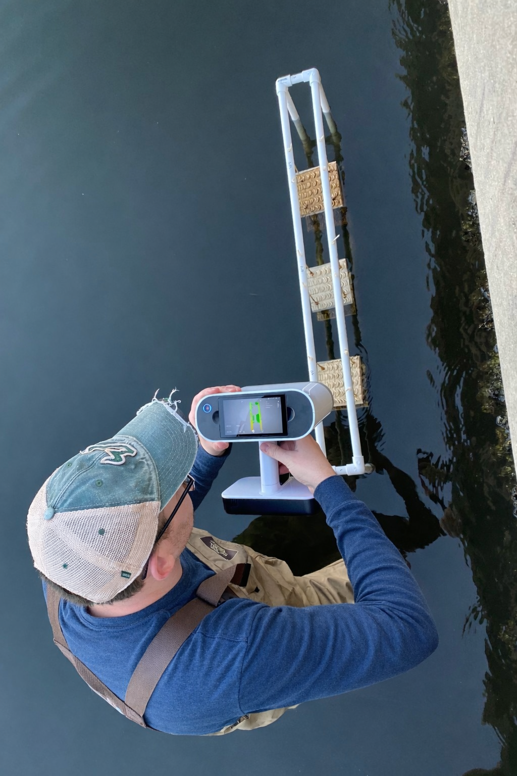 A person wearing a cap, glasses, blue long-sleeve shirt, and waders is standing by the water's edge, holding an Artec Leo 3D scanner device over the water, with a dock and water in the background.