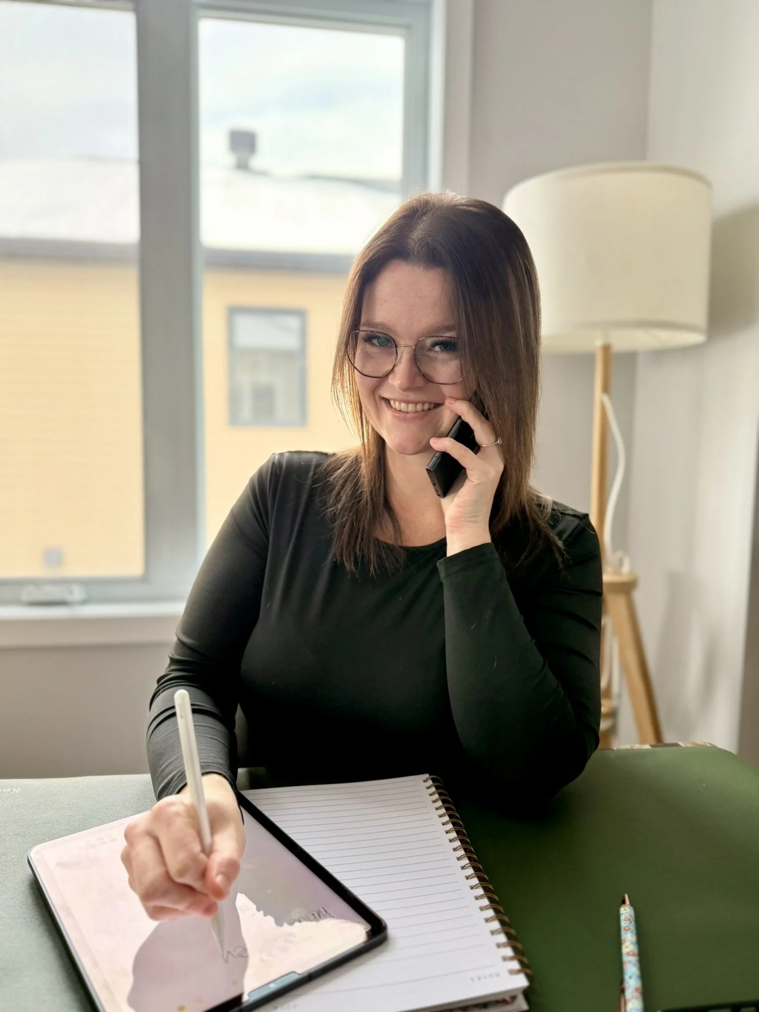 Femme souriante avec lunettes, assise à un bureau, parlant au téléphone, écrivant sur une tablette avec stylet, un cahier à lignes et un stylo coloré sur la table, dans une pièce lumineuse avec une grande fenêtre et une lampe à pied en arrière-plan.