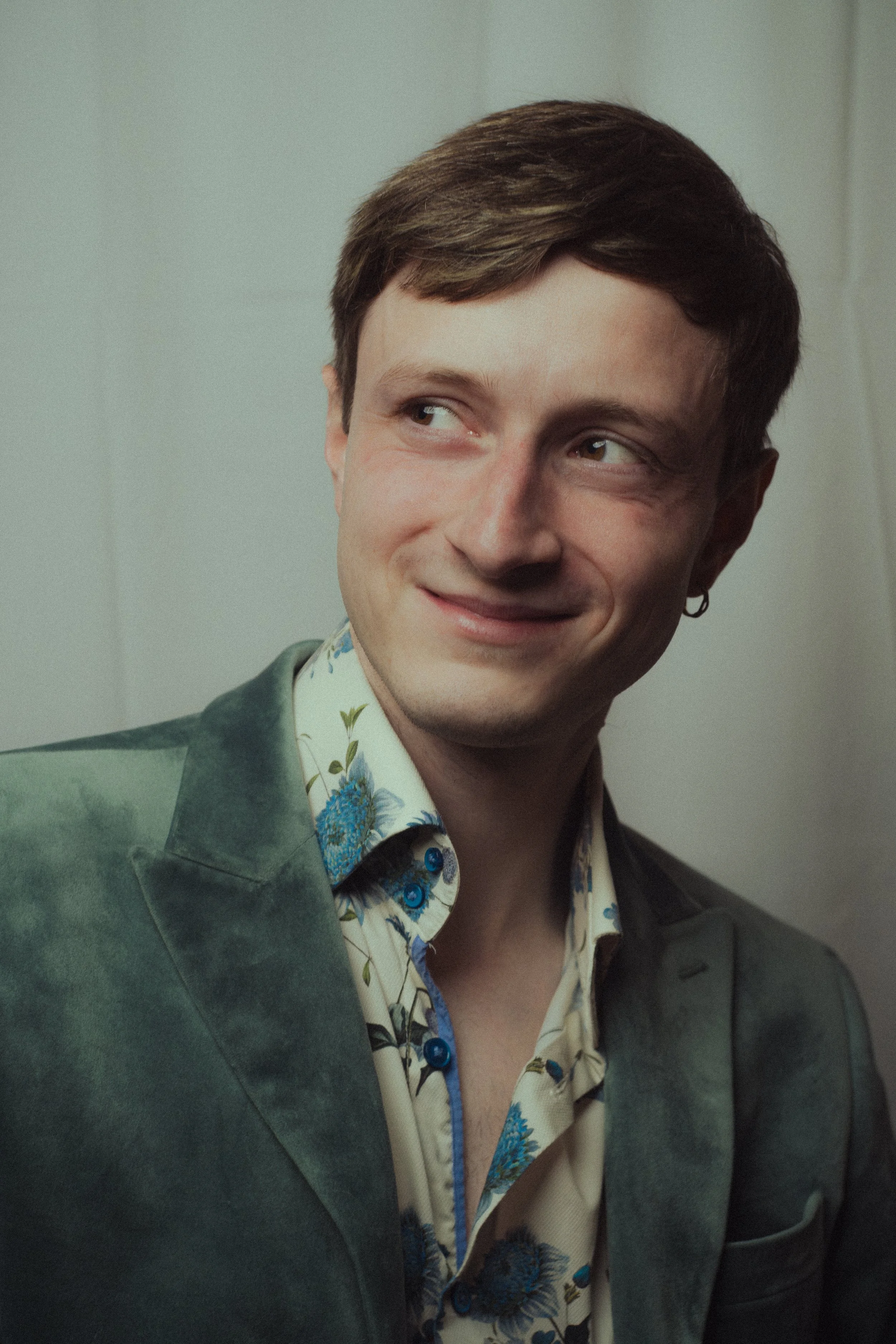 A man with brown hair, wearing a floral shirt and a green blazer, smiling and looking to his right.