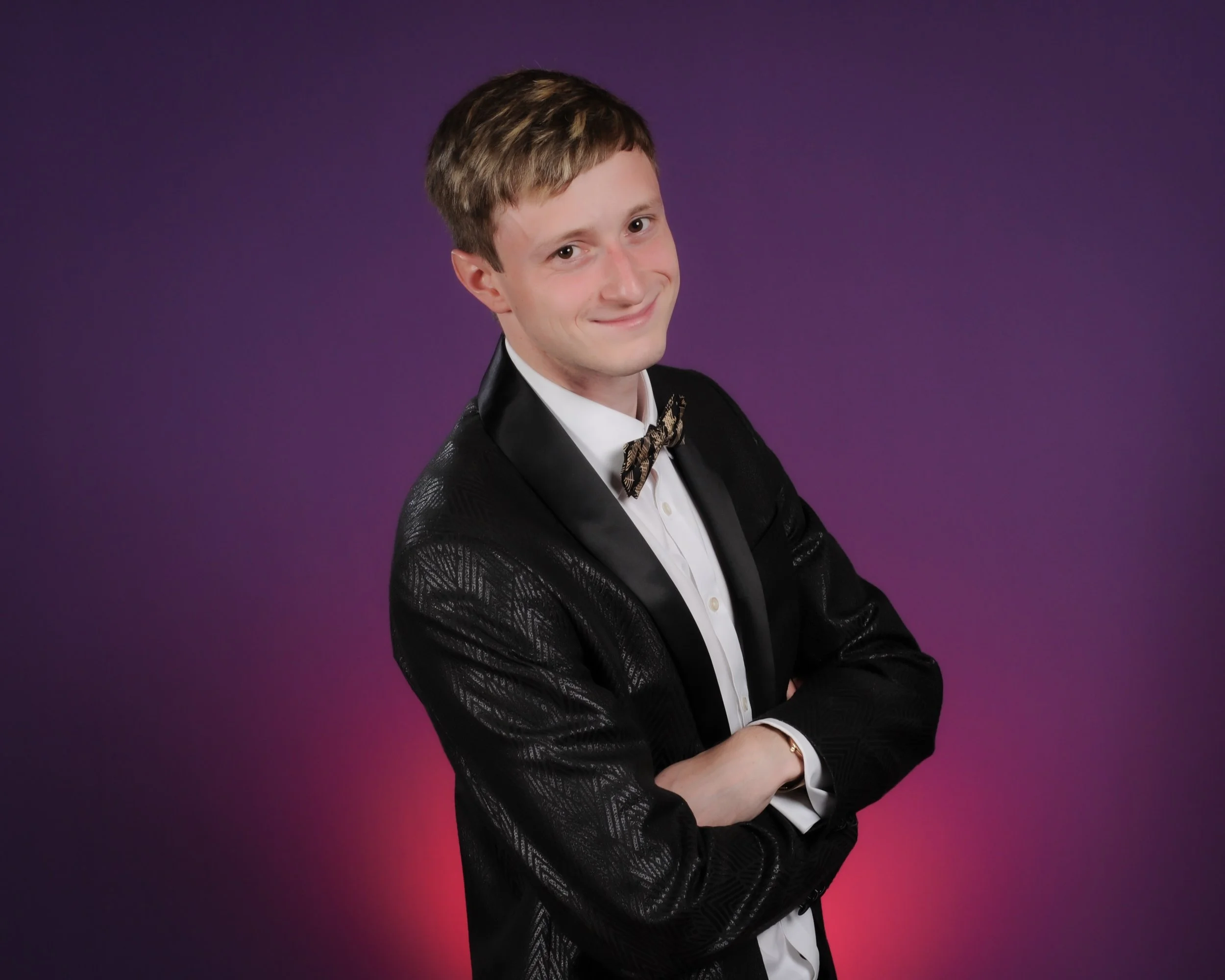 A young man with light brown hair, wearing a tuxedo with a patterned black jacket, white shirt, and a bow tie, standing with arms crossed against a purple gradient background, smiling gently.