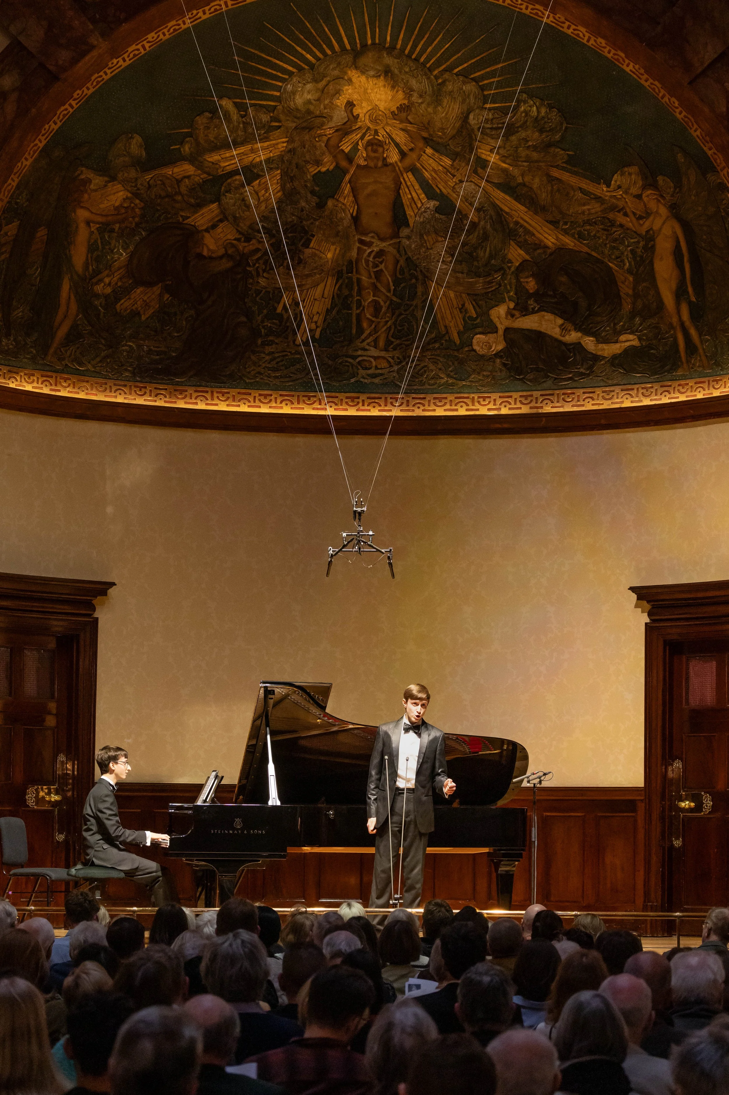 A classical music performance in a concert hall. A pianist is seated at a grand piano and a singer is standing, performing. The audience is watching attentively. An ornate ceiling mural with religious imagery is visible above the stage.