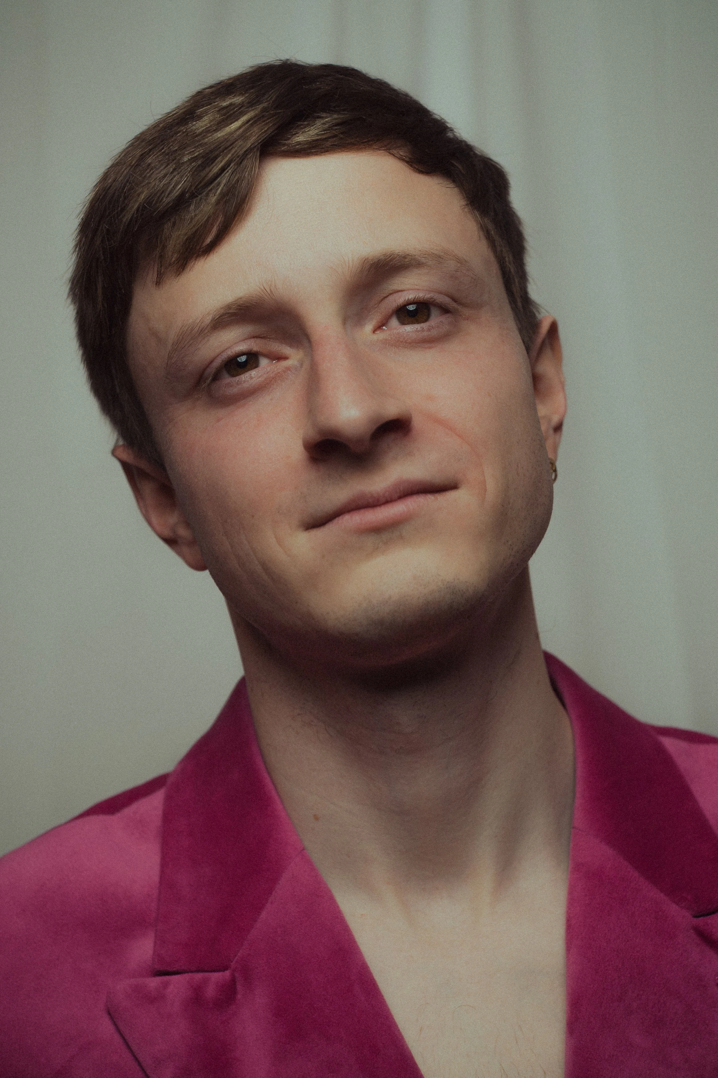A young man with short brown hair and a slight smile, wearing a pink satin blazer, standing against a plain background.