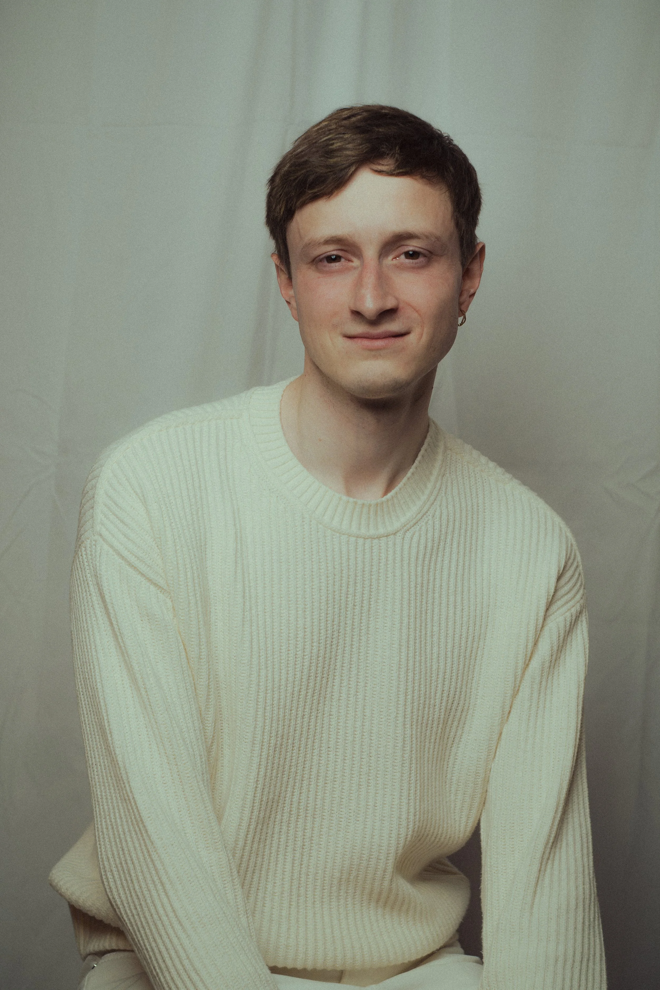 A young man wearing a cream-colored, ribbed sweater, sitting against a pale background, facing slightly to the right with a soft smile.