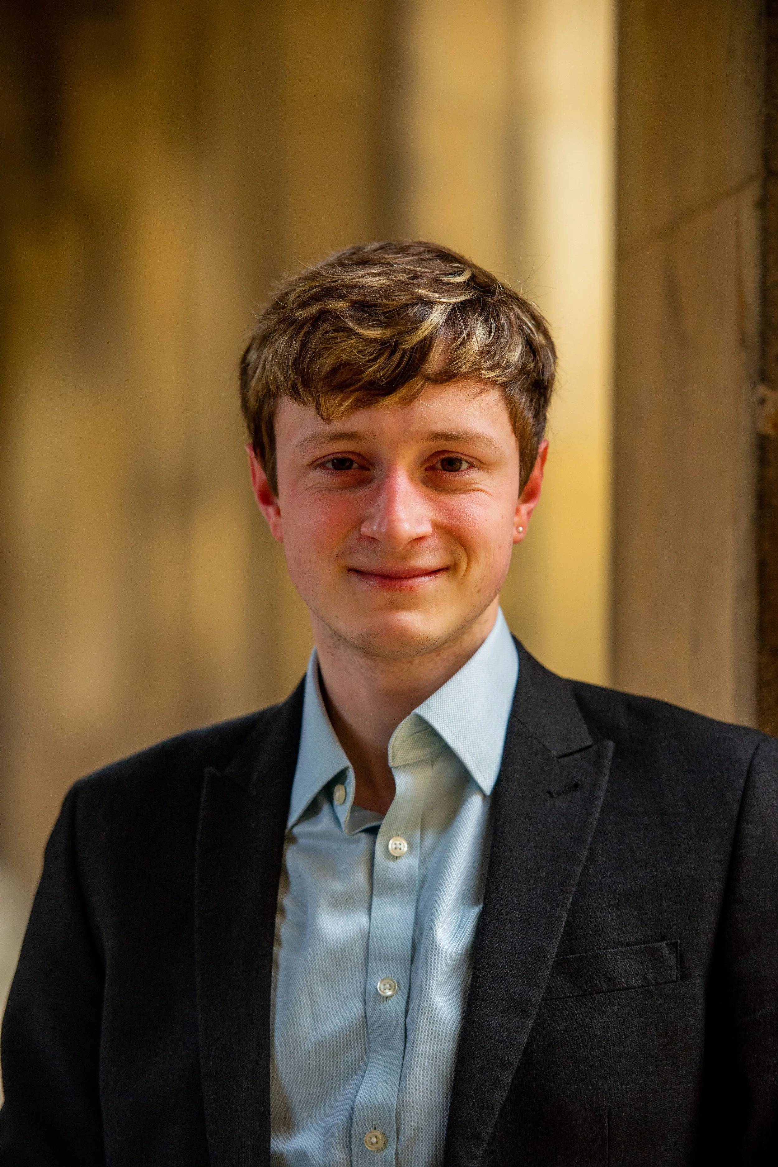 A young man with light brown hair wearing a black blazer and light blue dress shirt, standing in front of a blurred background with warm yellow lights.