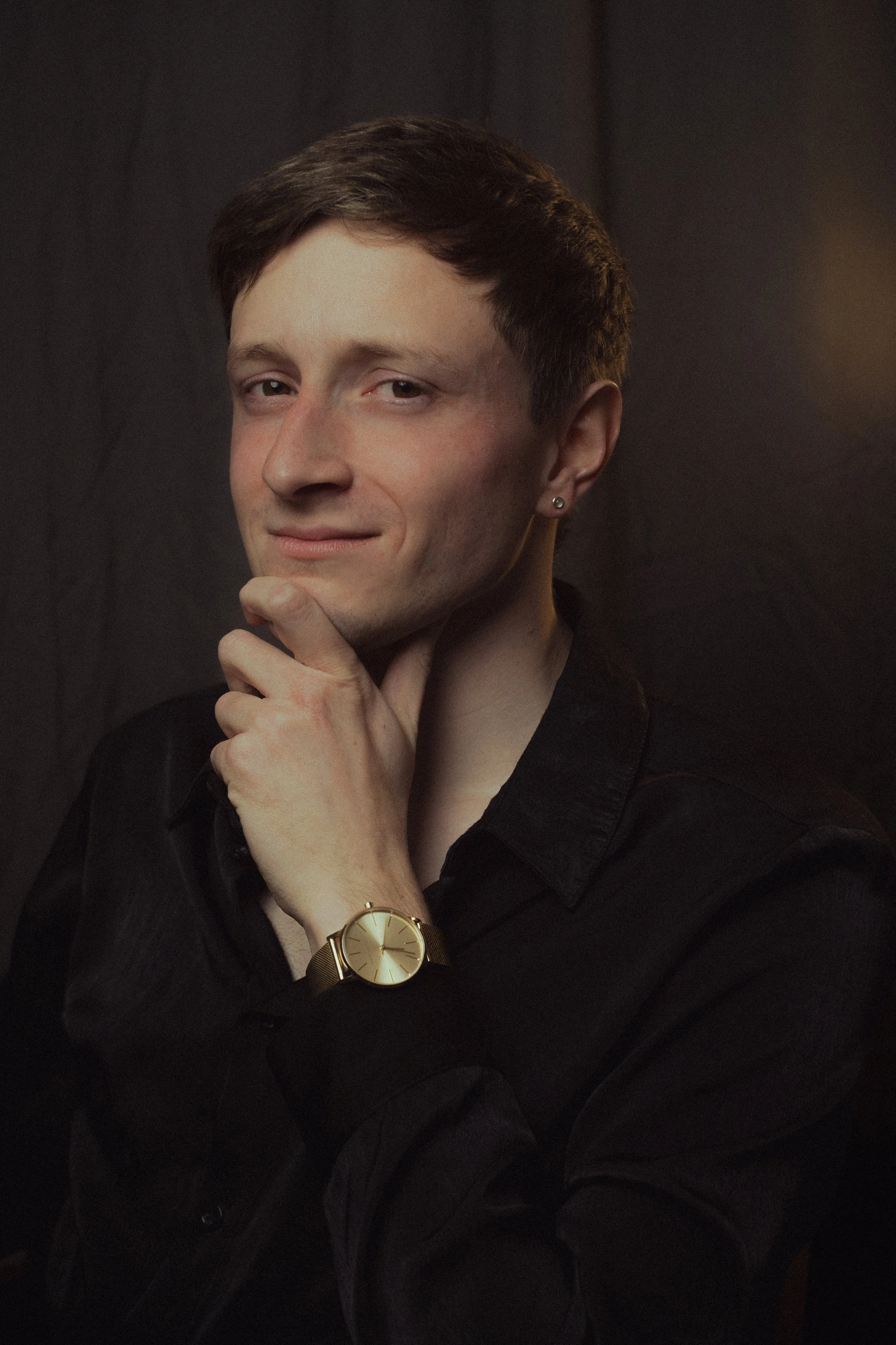 A young man with short brown hair, wearing a black shirt and a gold watch, resting his chin on his hand with a slight smile, in front of a dark background.