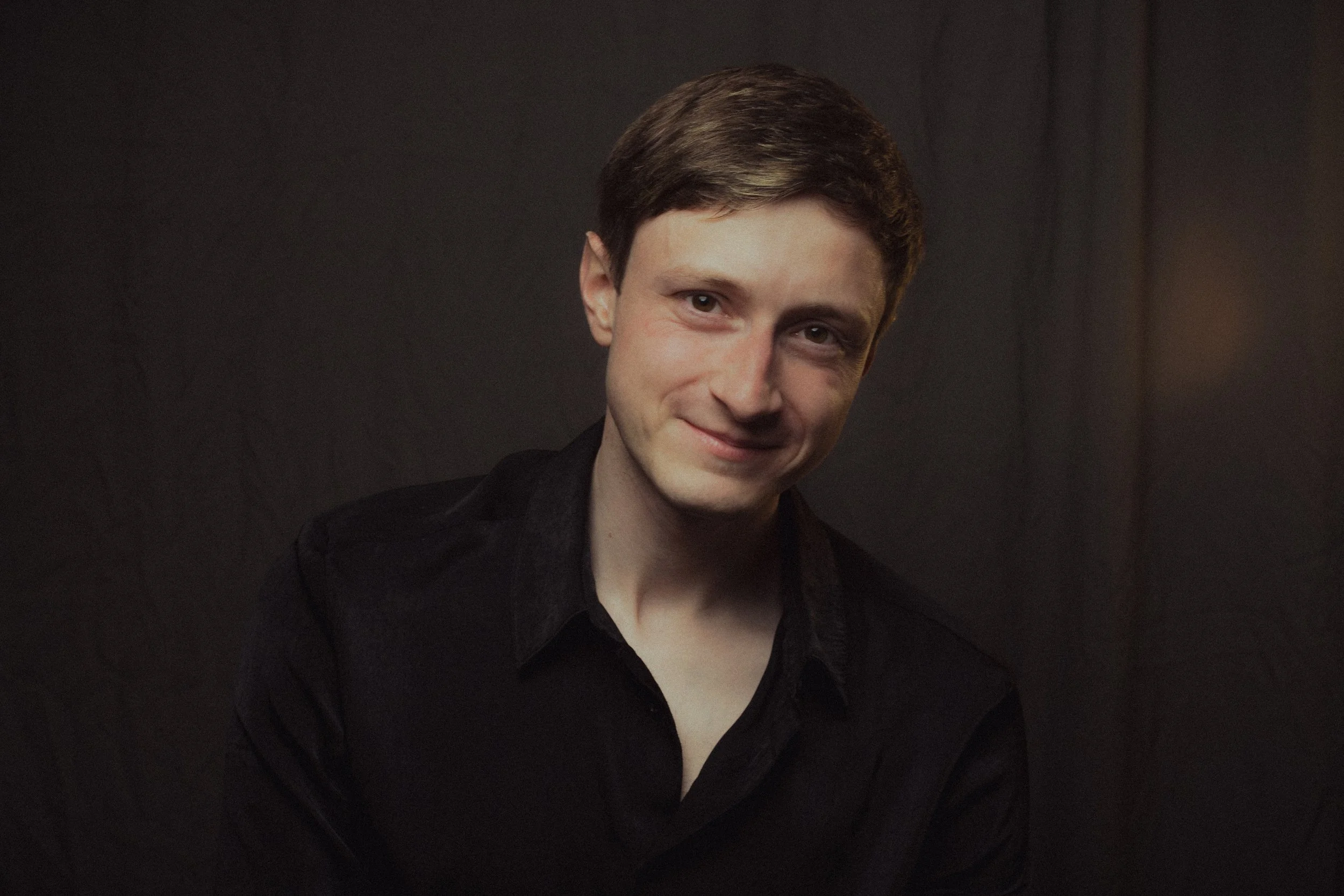 A young man with short brown hair smiling, wearing a black shirt, against a dark background.