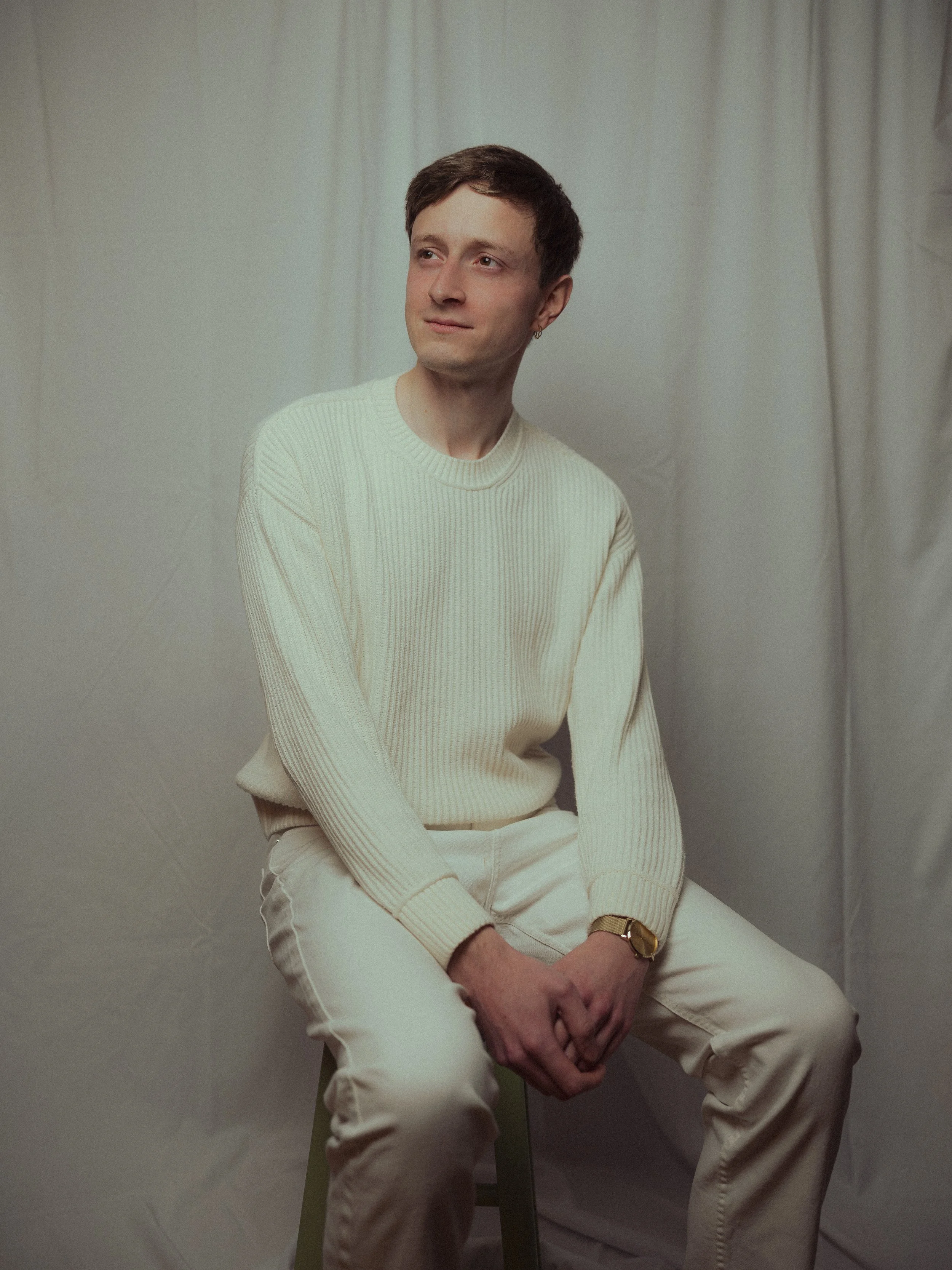 A young man with short dark hair sitting on a stool against a neutral background, wearing a cream sweater, cream pants, and a gold watch, looking slightly upwards with a thoughtful expression.