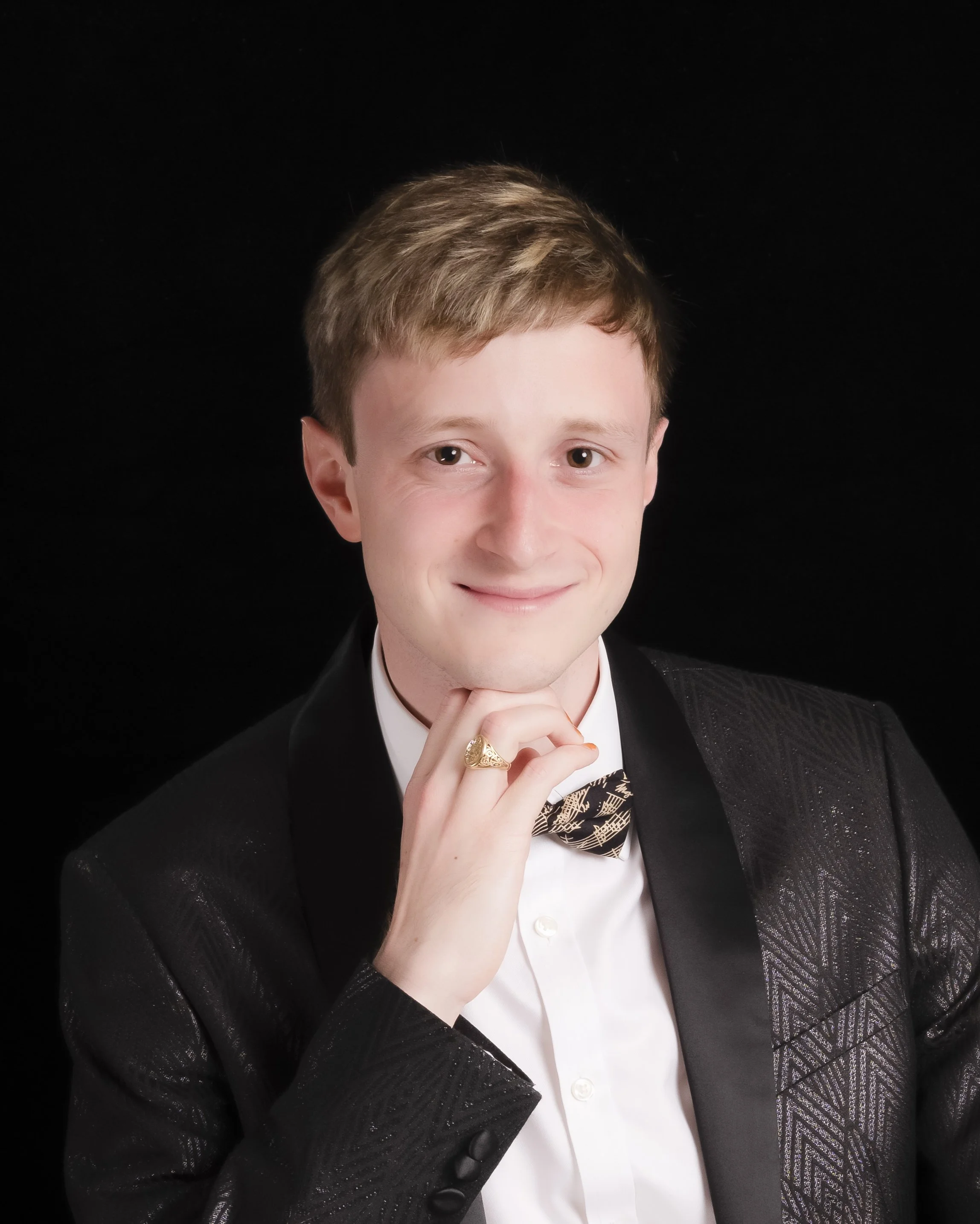 A young man with light brown hair in formal attire, wearing a black tuxedo with a patterned bow tie, against a black background, with his hand resting under his chin and smiling.