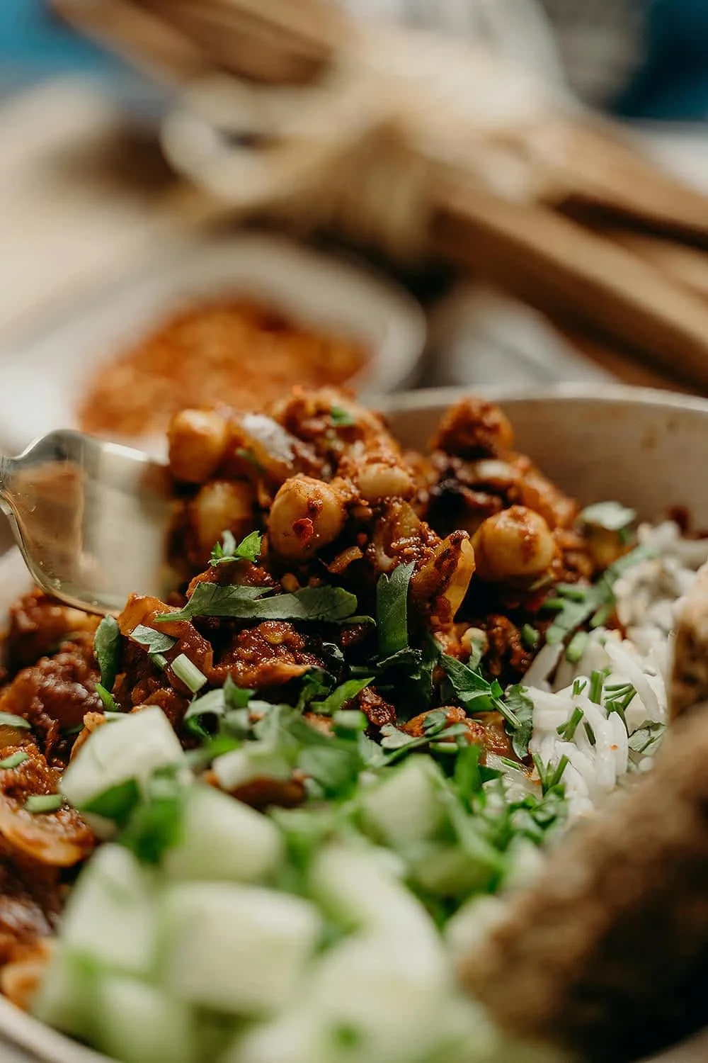 Close-up of a dish of chickpeas and ground beef with chopped green onions and herbs, served with flatbread on a table with small bowls of spices and utensils in the background.