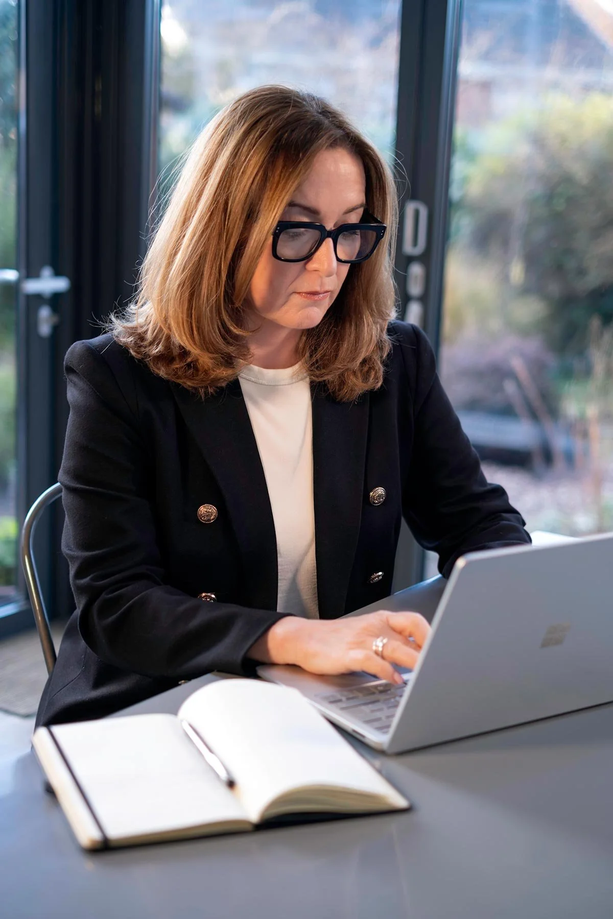 A woman with shoulder-length brown hair, wearing glasses, a black blazer, and a white top, is working on a laptop at a table in a room with large windows showing a blurred outdoor background.