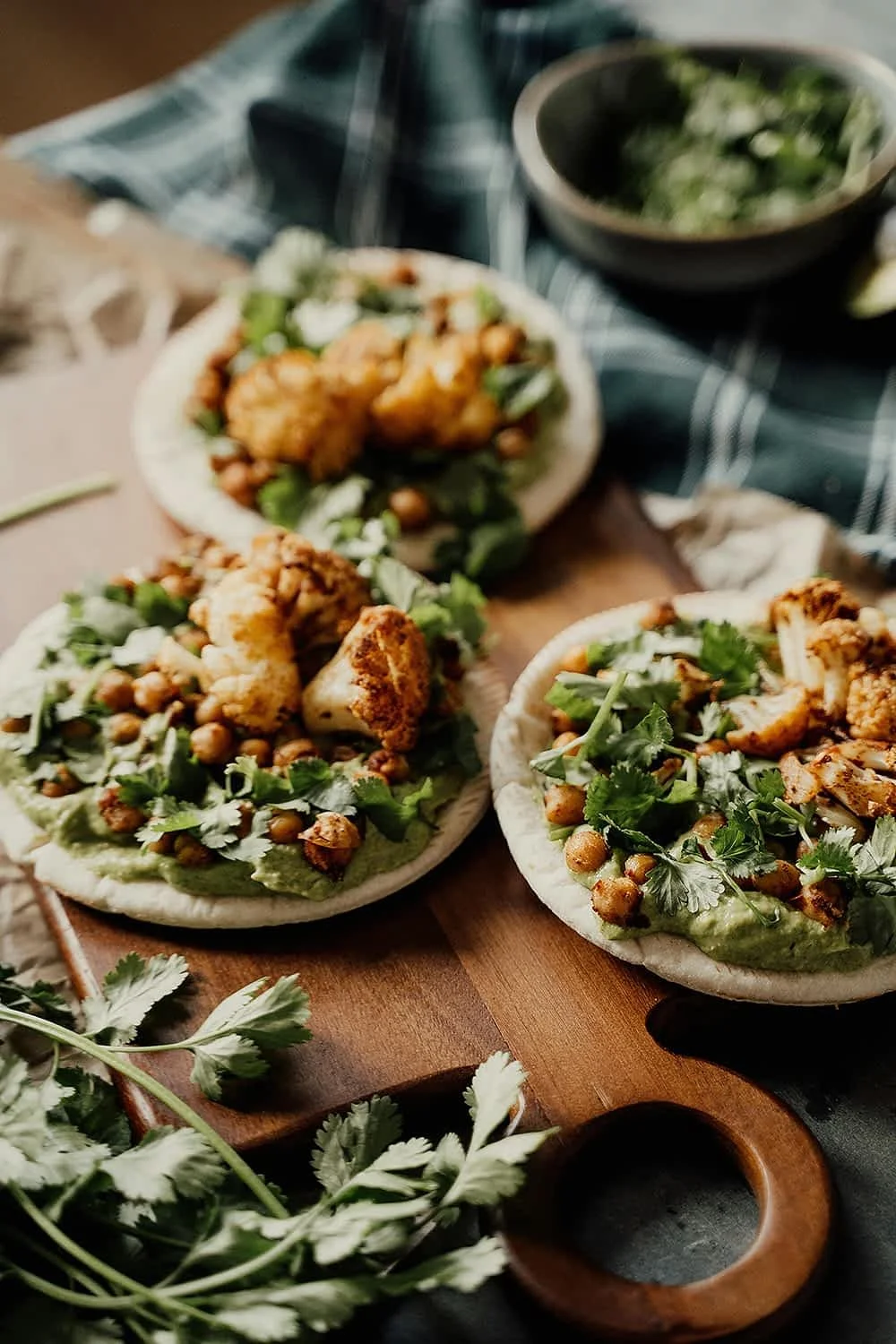 Three pita pizzas topped with roasted chickpeas, cauliflower, fresh cilantro, and a green sauce, set on a wooden board with a bowl of salad in the background.