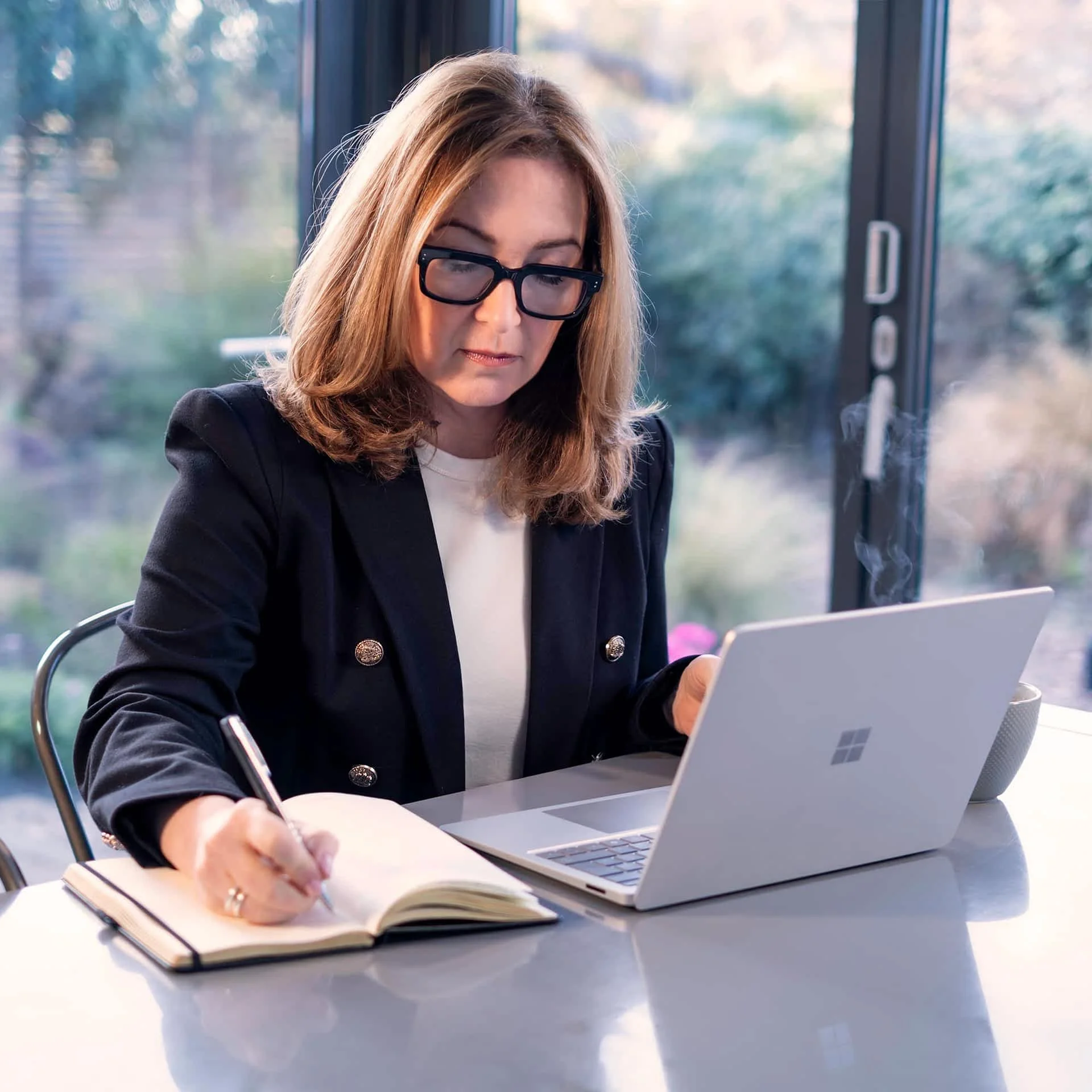 A woman with shoulder-length hair and glasses sitting at a table, writing in a notebook with a pen, with a laptop open in front of her, in a setting with large windows showing trees outside.