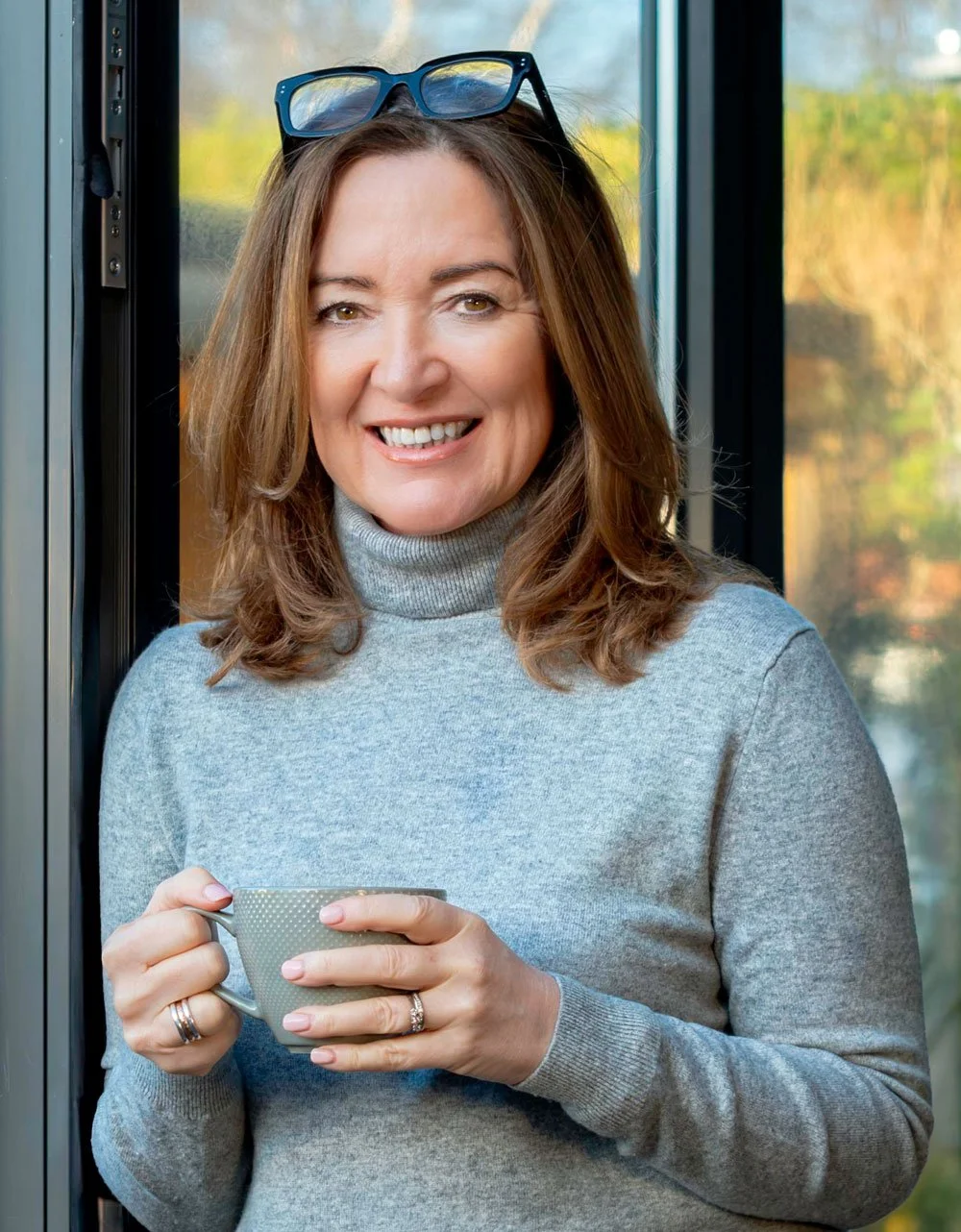 A woman with glasses on her head smiling, holding a mug, standing near a window with a blurry outdoor background.