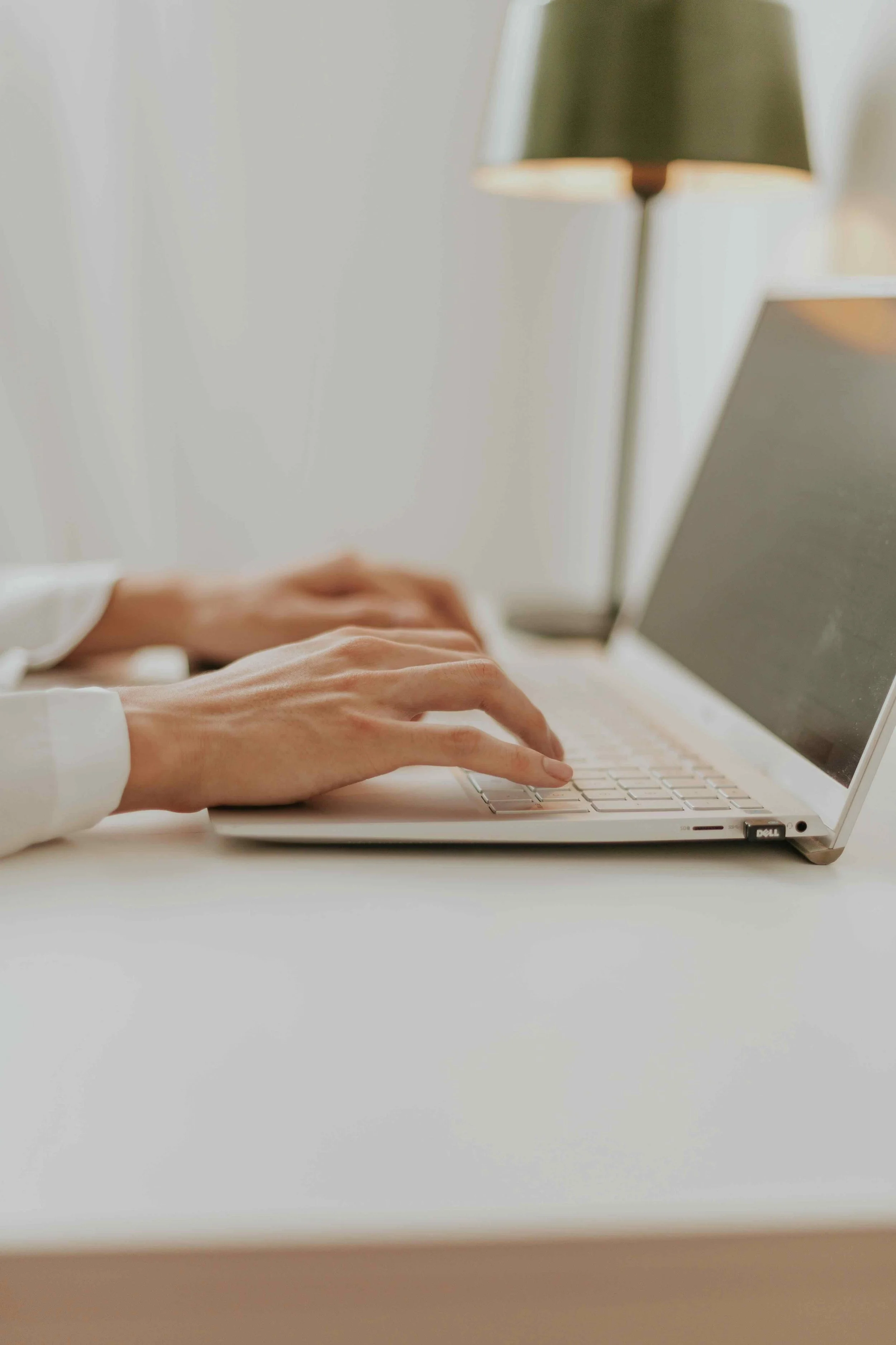 Close-up of hands typing on a laptop keyboard at a desk with a softly lit lamp in the background.