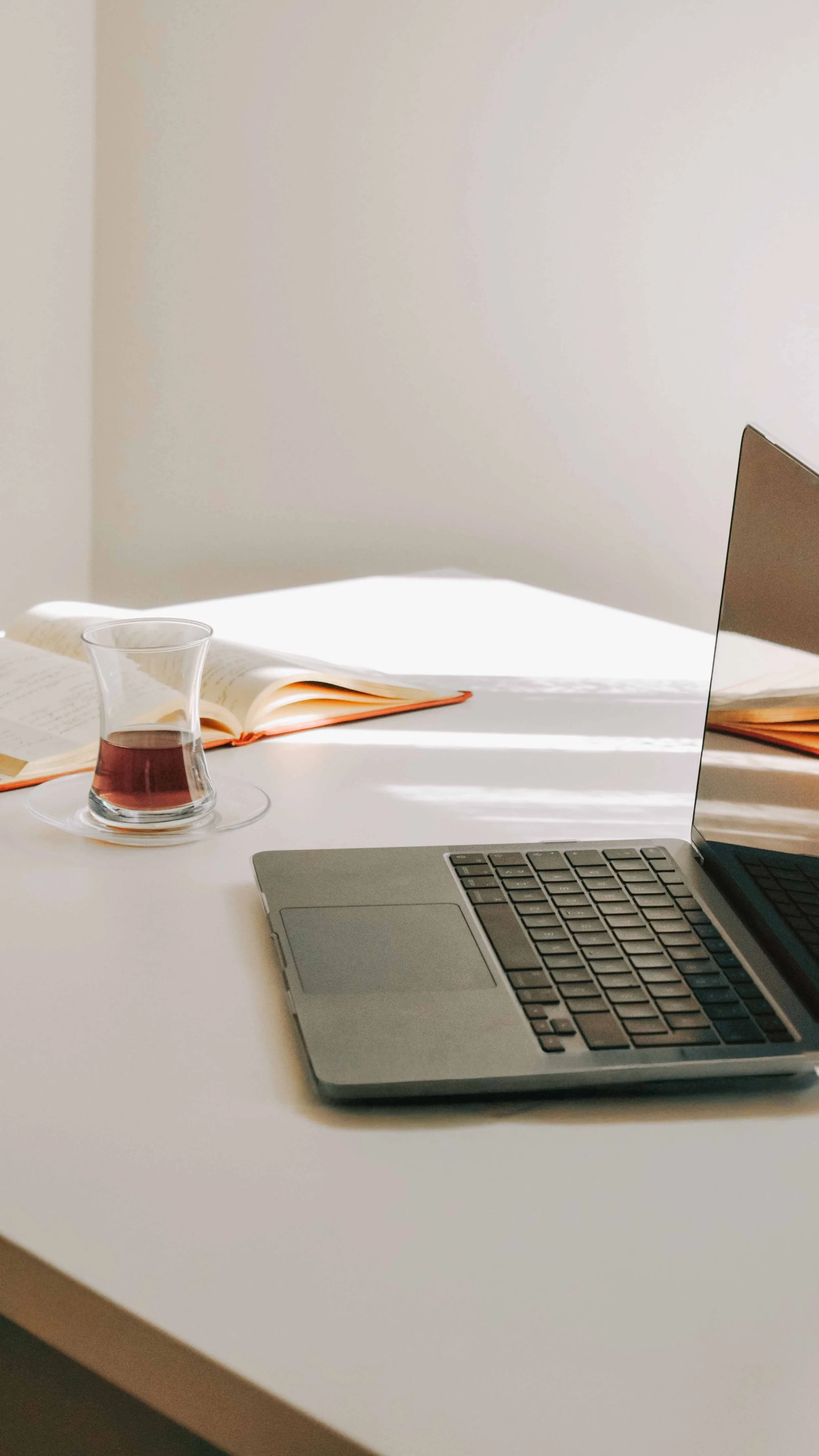 Open laptop on a white desk beside a glass of tea and an open book in warm natural light.