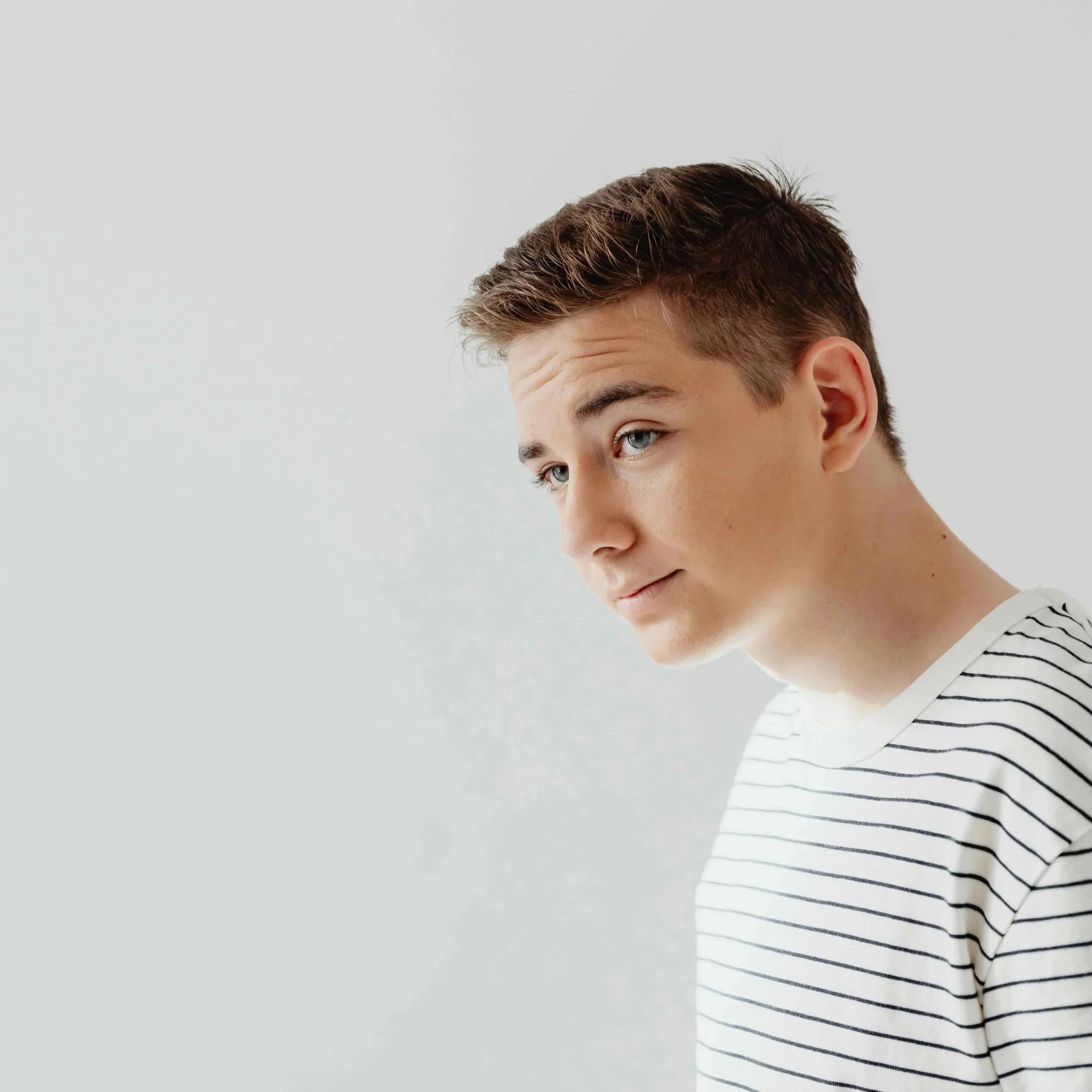 A young man with short brown hair and blue eyes, wearing a white and black striped shirt, looking slightly downward and to the side against a plain white background.